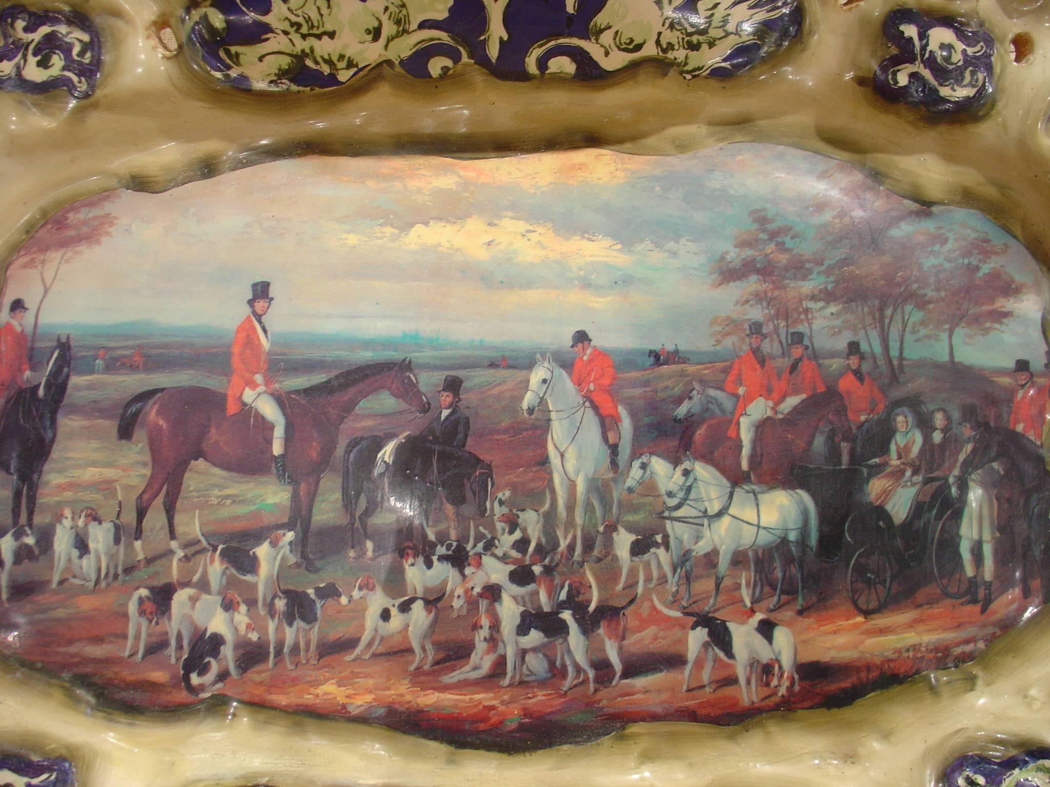 This antique ceramic platter has a central hand-painted hunting scene and originates from Northern Brittany. There is an irregular border surrounding the scene that has insets of raised hand-painted sections. The scene is that of a Classic English