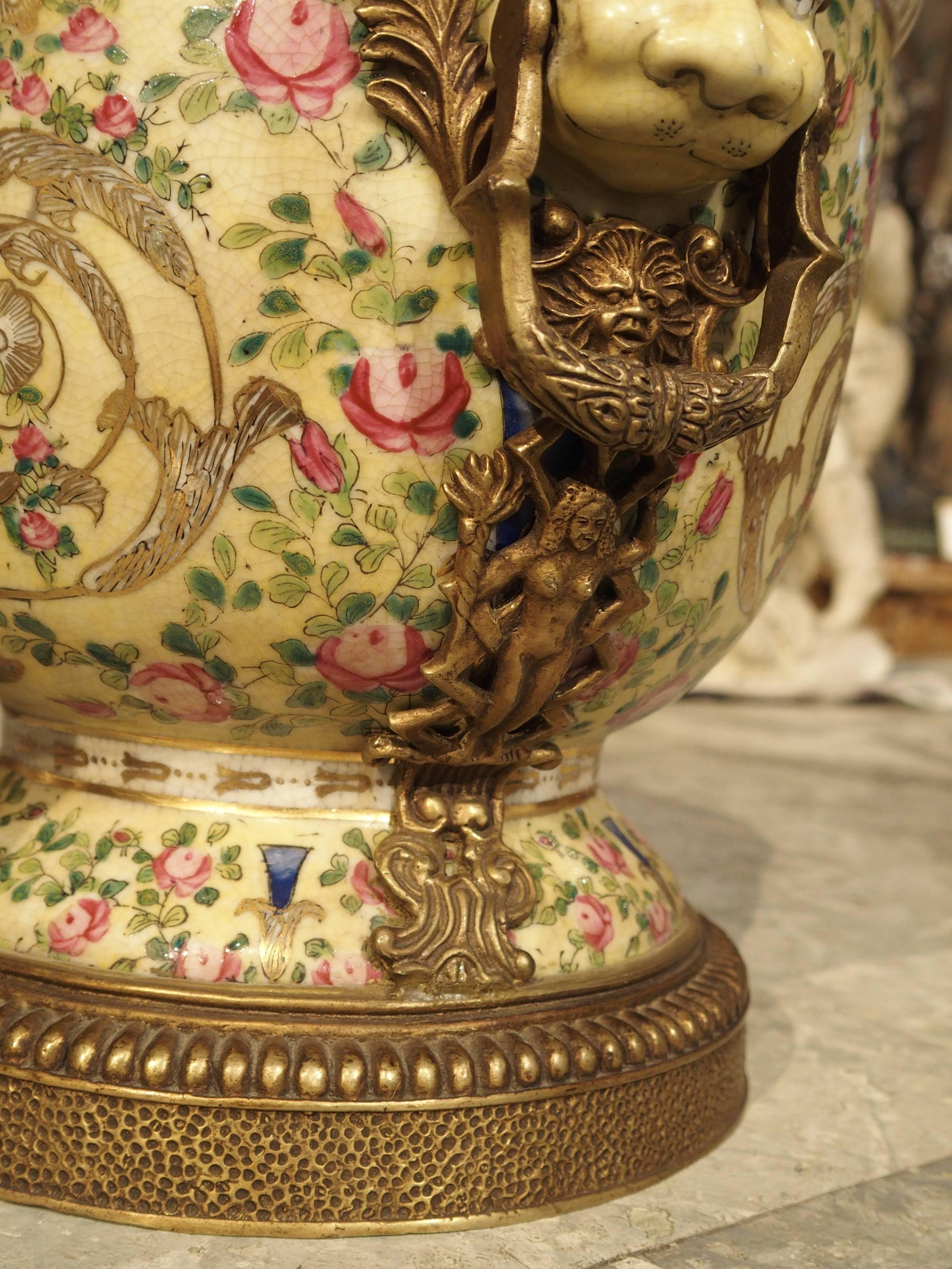 This elegant pair of painted porcelain urns has gilt metal mounts surrounding the top, the base, and the trim surrounding the metal handles that extend from the top rim down to the base. The raised painted motifs are of scrolling pink cabbage rose