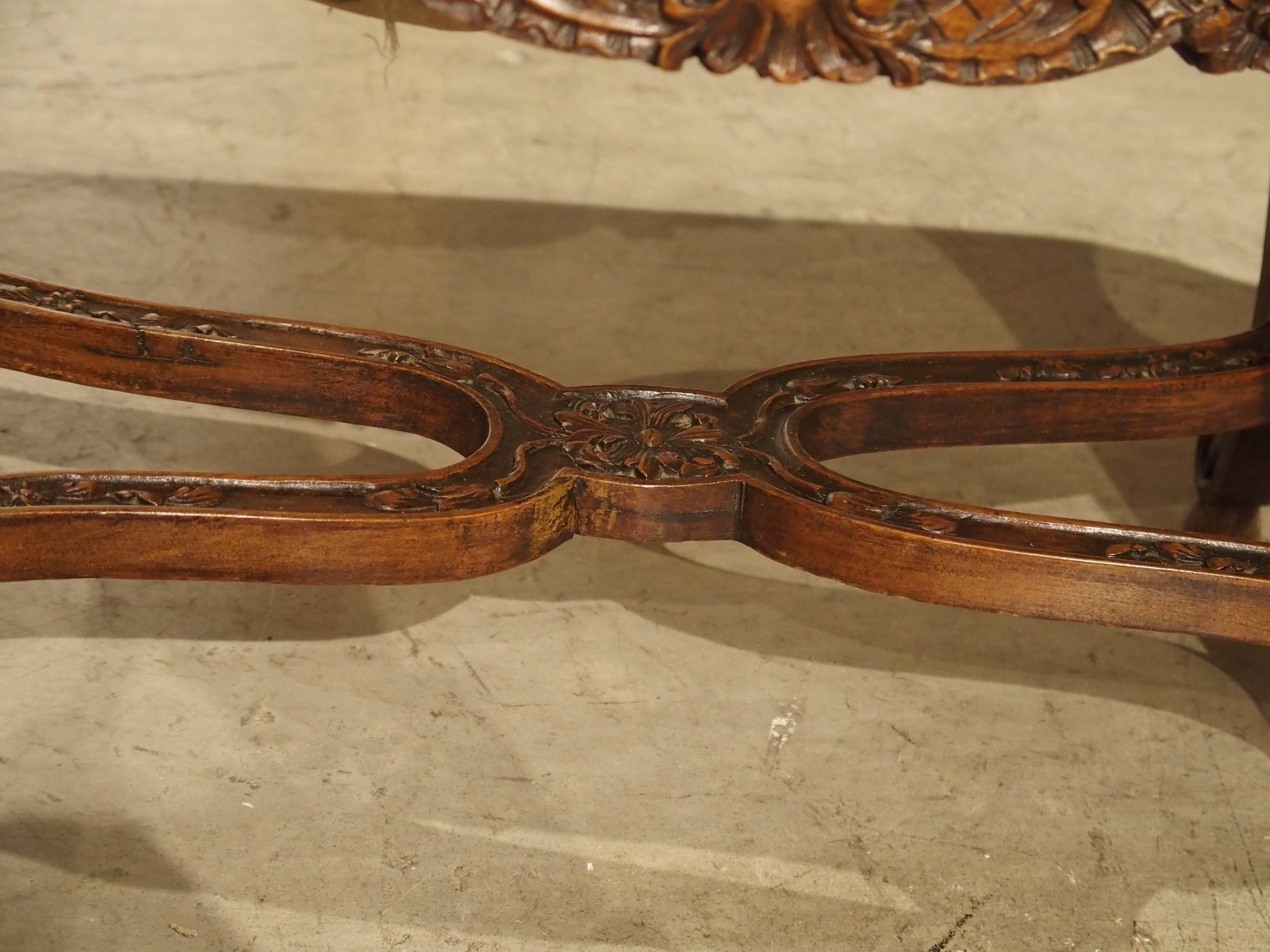 This magnificently carved antique French upholstered bench has a shaped apron along all sides. The top has been upholstered many years ago in a fine silk brocade. It is attached with brass nailheads. There are six shaped and carved legs with finely