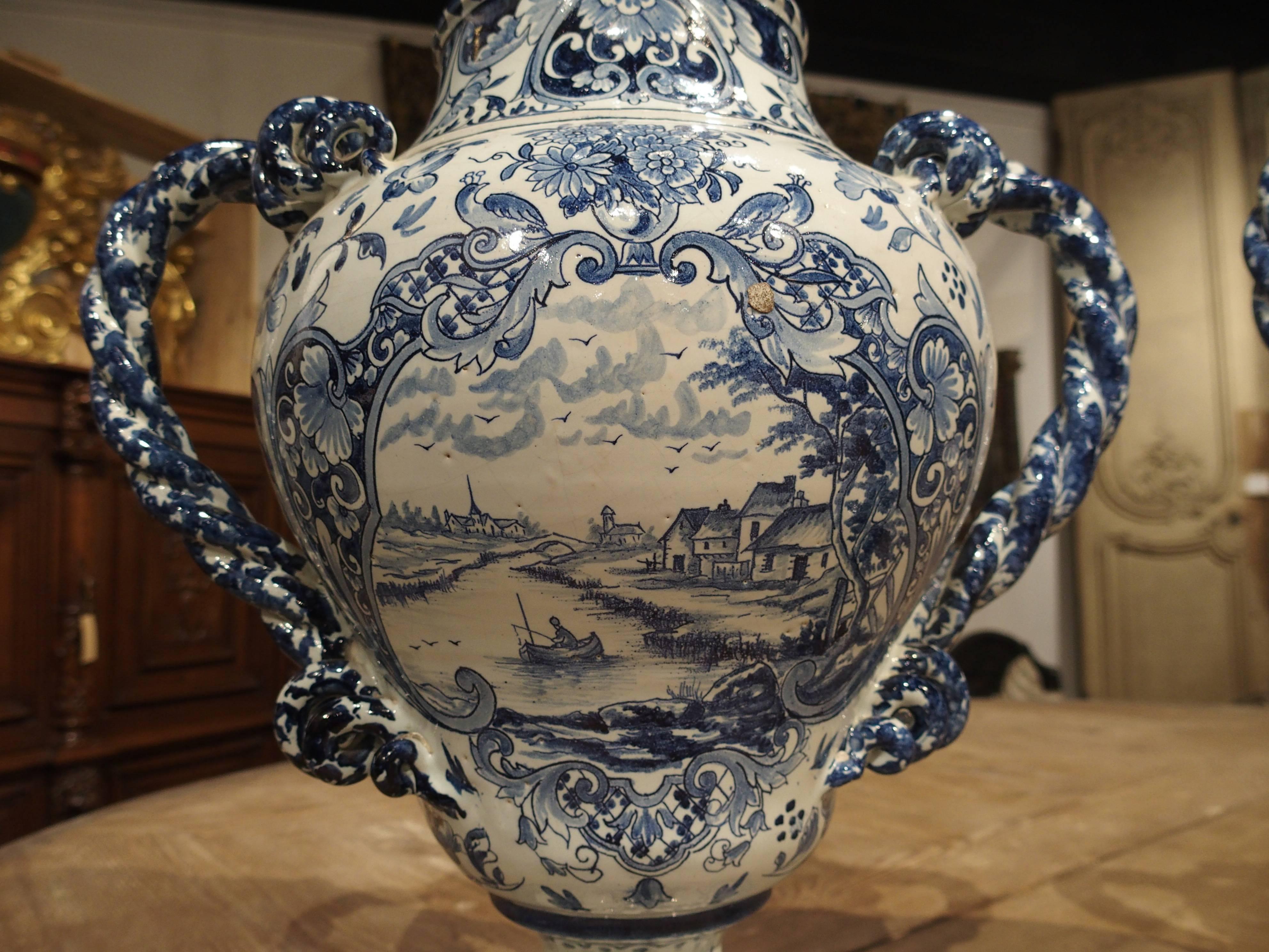 This beautiful pair of scenic blue and white vases have twisted rope handles to either side. Both sides of each depict central scenes within cartouches which have been hand-painted. On one side of each is a different scene depicting sailboats