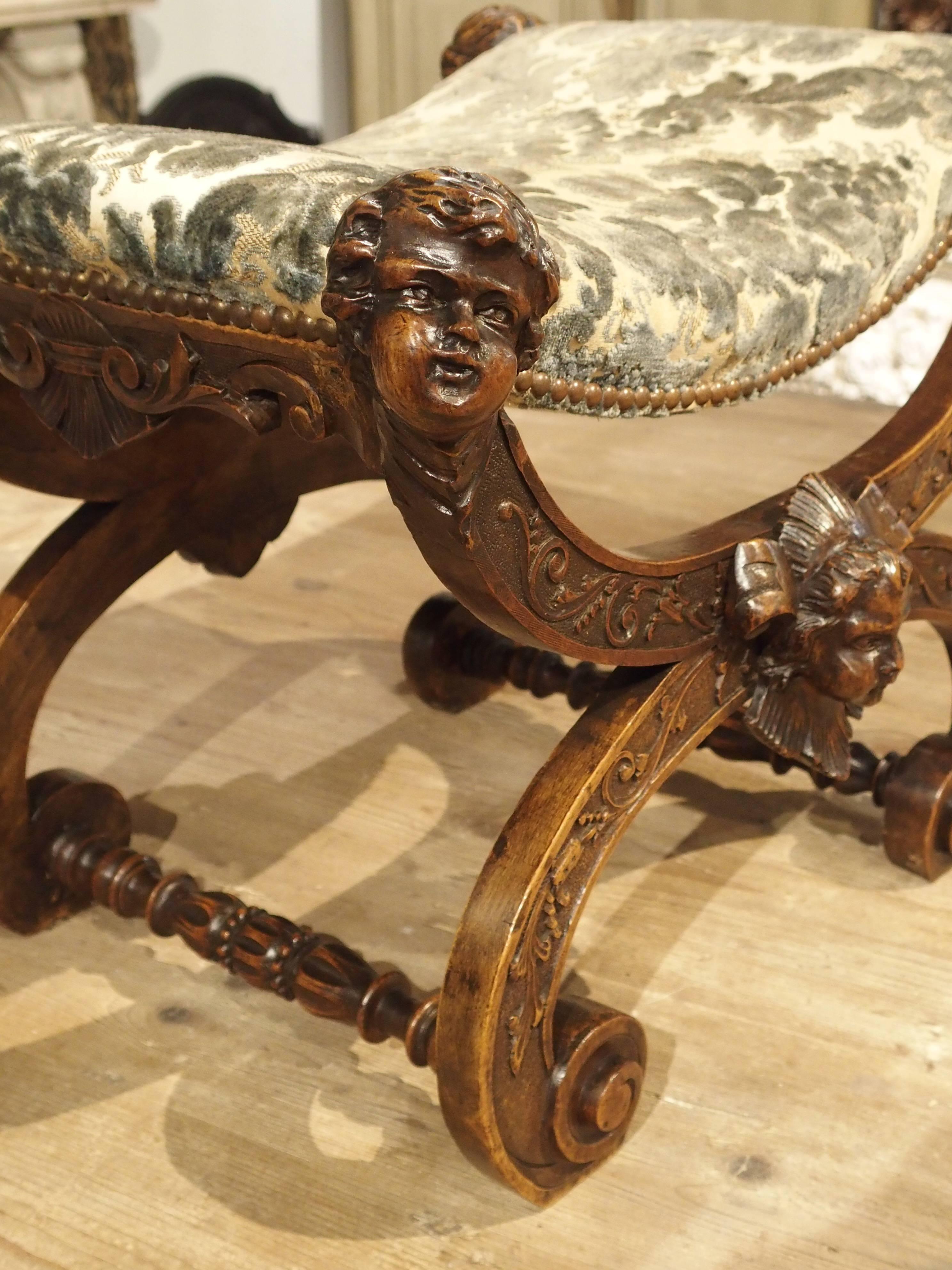 This elegant antique Italian stool is made of walnut and has X-framed legs with turned feet. The pleasant faces on all corners are uplifted and turned. On the legs, are scrolling floral and foliate vines. At the centre points of the legs are more