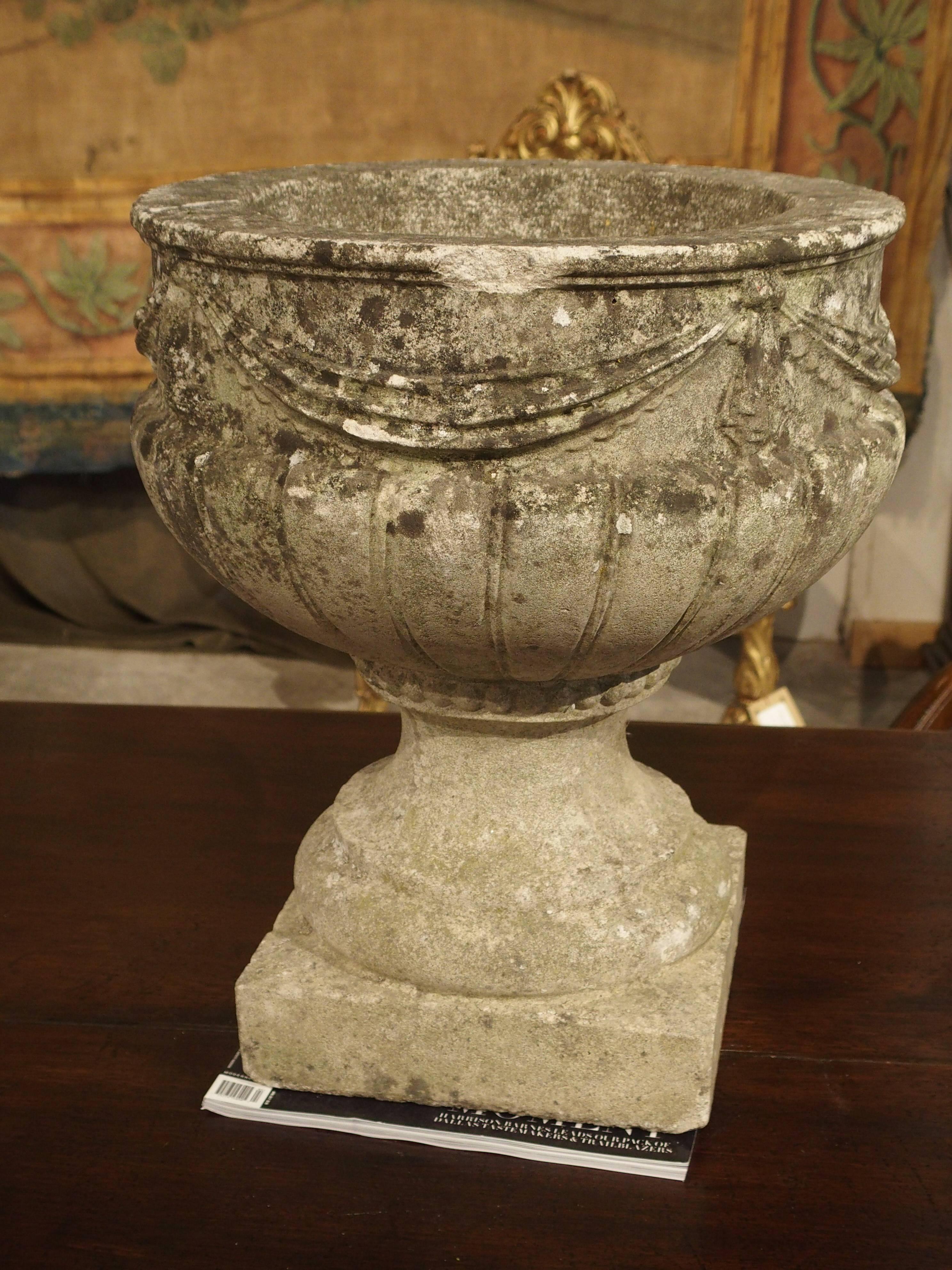 This planter from Belgium has the look of a very old solid limestone planter. It has drapery swags at the frieze and stylized gadrooned lobes beneath. It rests upon a square base. The edges are soft and worn. This beautiful vintage cast stone