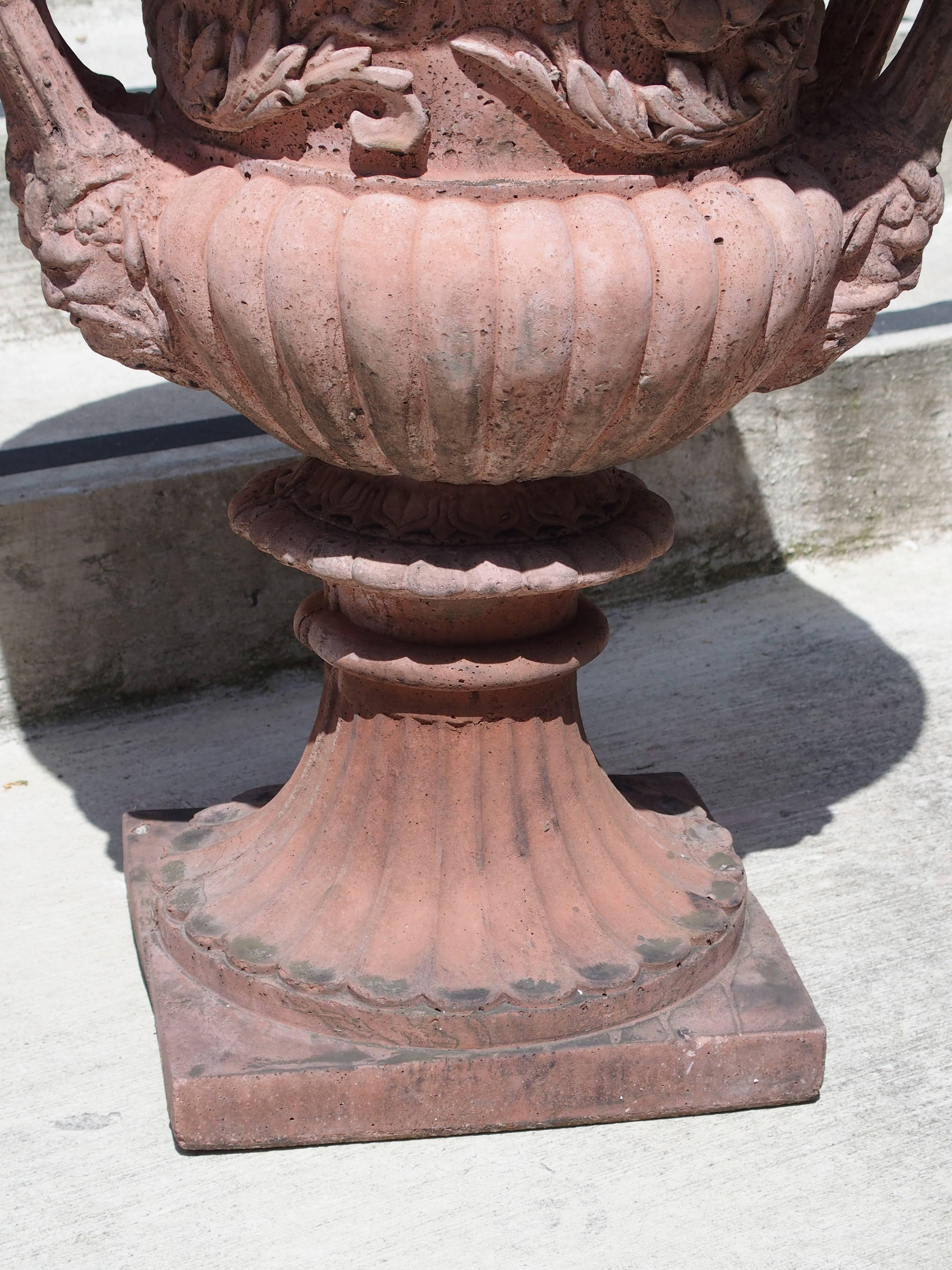 These cast stone urns have been made with a mixture of crushed limestone, bluestone, and cement. The terracotta color has been mixed in with the stone, not applied. Each urn has two handles with double mascarons. Gadrooned lobes are beneath, which