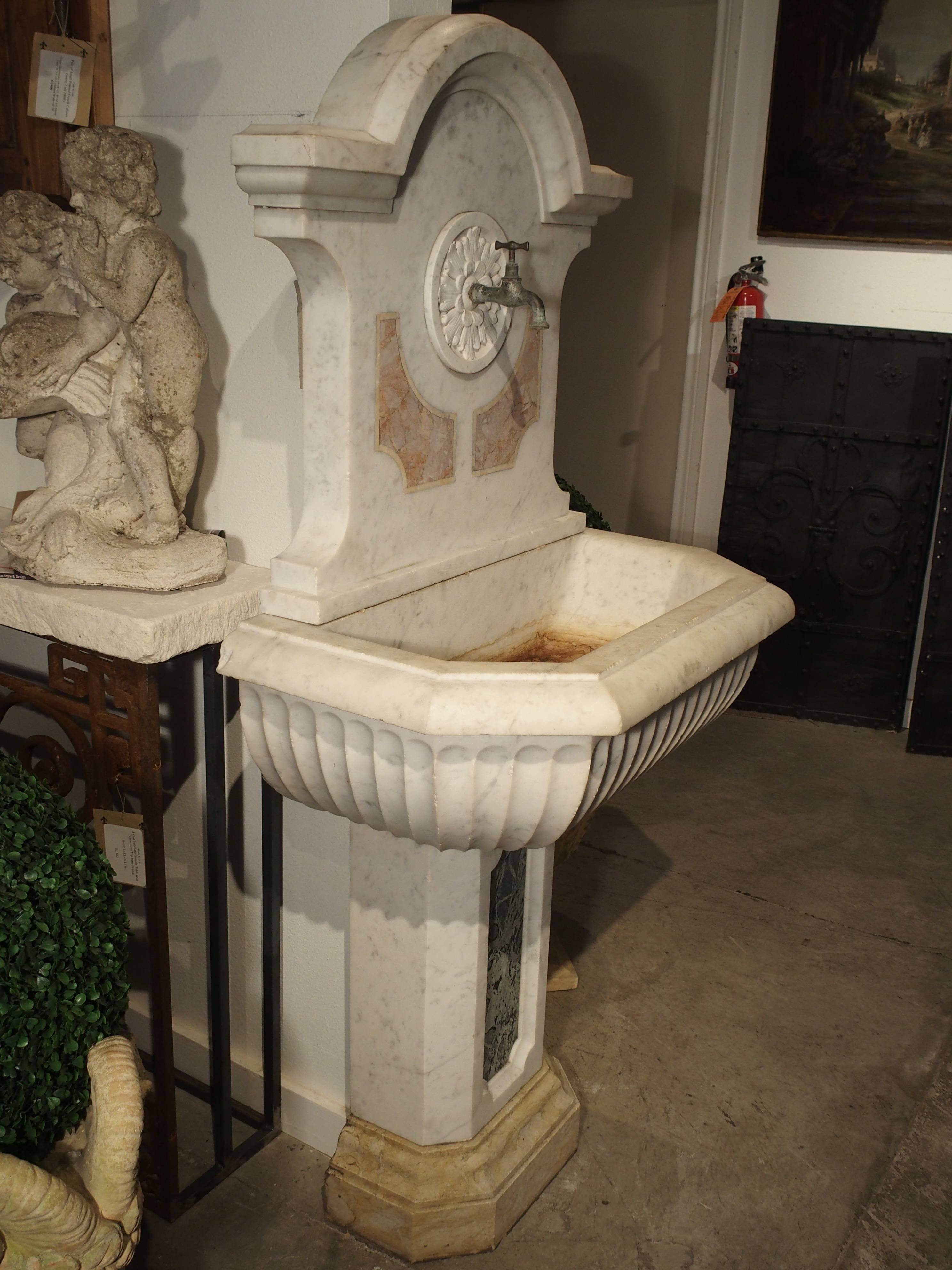 This elegant antique Carrera marble Italian wall fountain has an arched, stepped in top with right angles to the sides. Beneath this and on the back plate, there is a round circle filled with a stylized palmette and faucet for dispensing of the