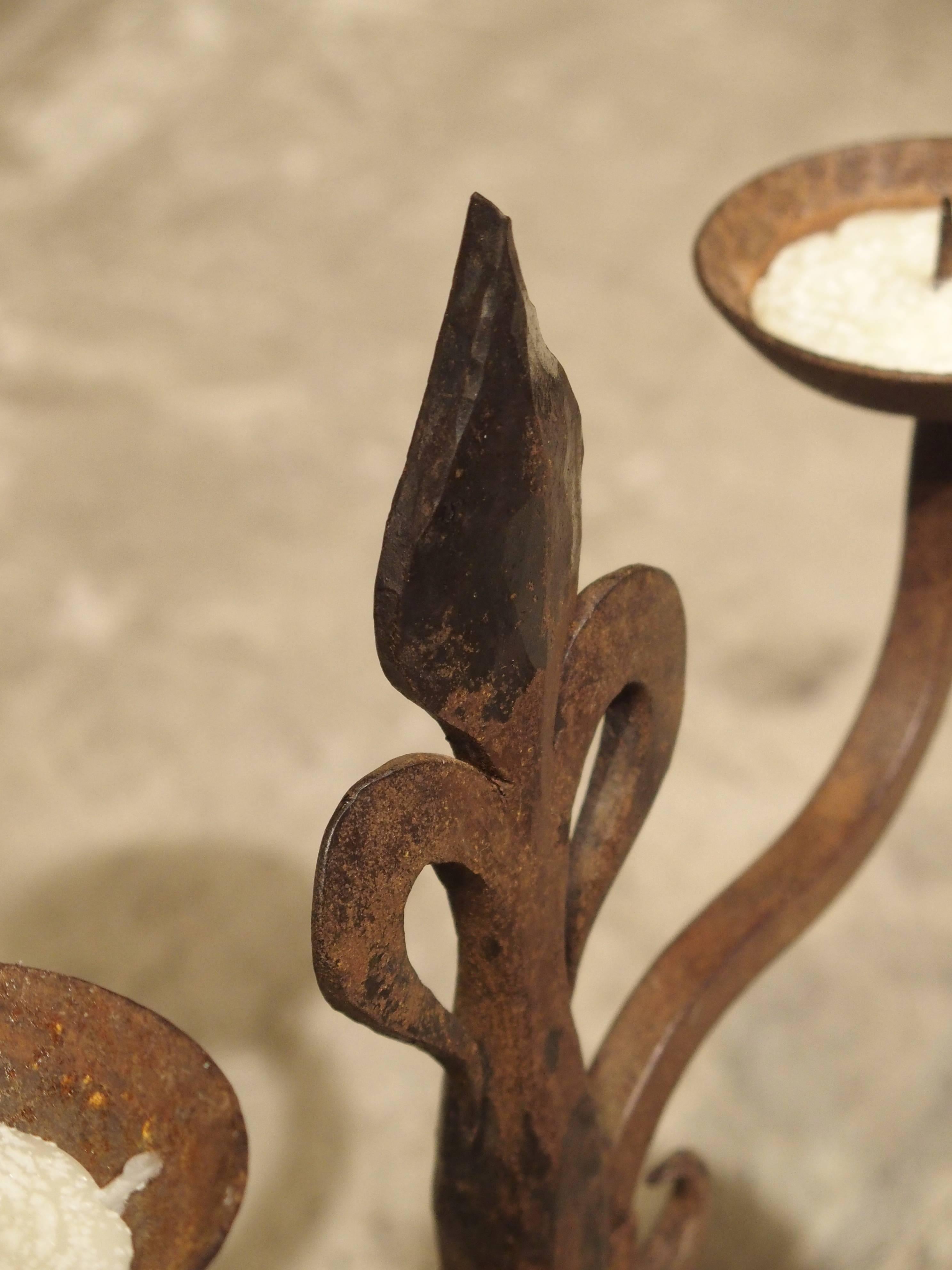 These two-arm French candelabras are hand-forged and hammered. There are two S-scroll arms banded to a center post with the Fleur de Lis at the top. This rests upon four legs placed in a rectangular shape. Perfect for any area of the home, these