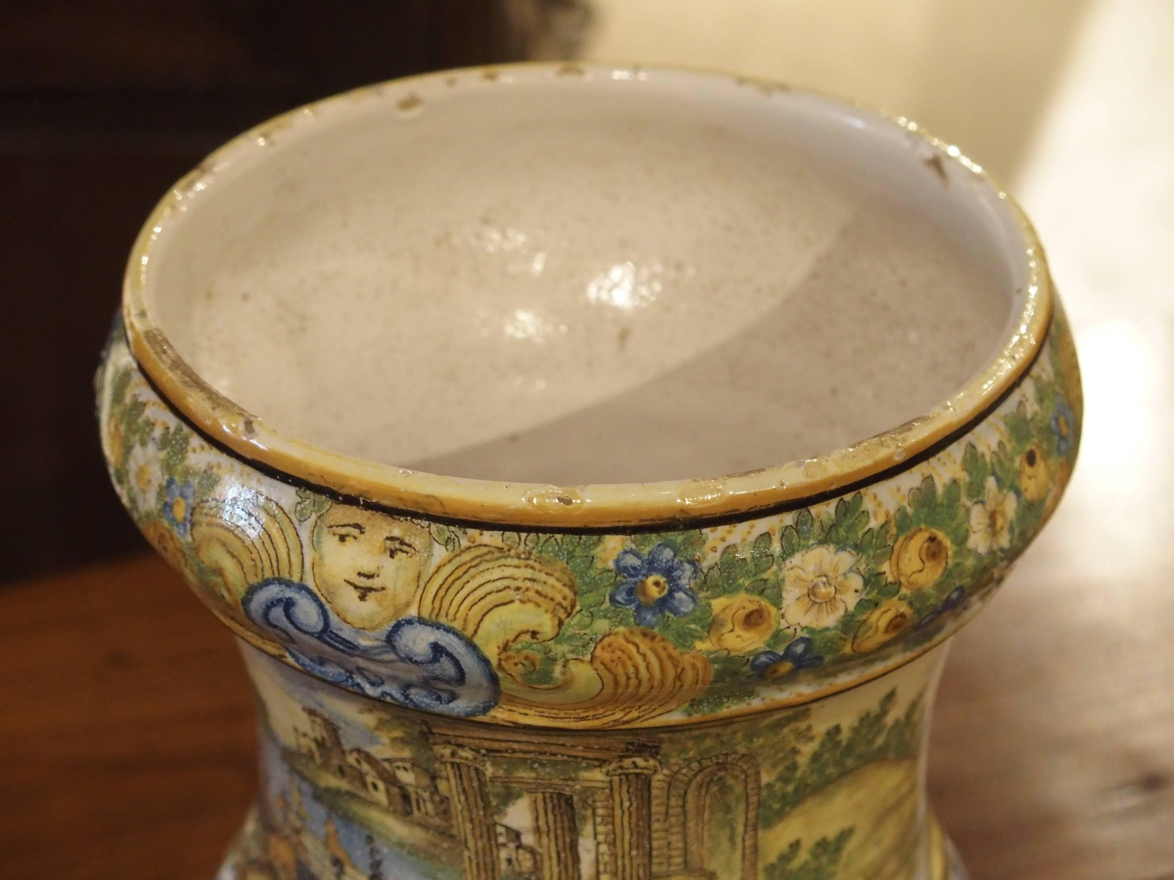This charming antique hand painted urn from Italy depicts a continuous, all around scene at its centre. The main colors are a mix of soft yellows, blues, and greens. The main subject is a large body of water with a variety of structures and figures