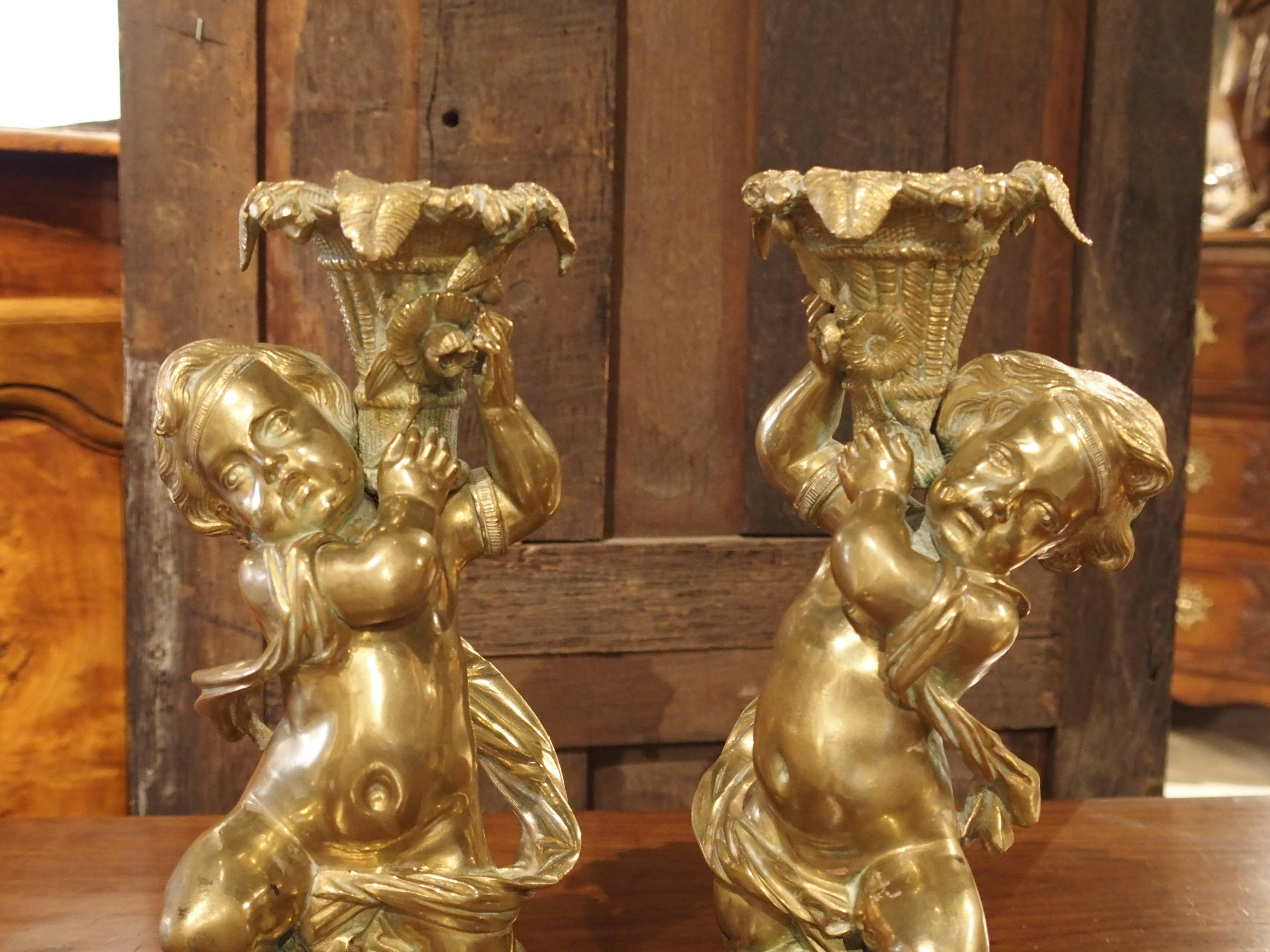 These charming gilt bronze candlestick holders are French and date to the mid-1800s. They are a refined quality and have a very nice color. Each depicts a well detailed putti on a foliate mound with a classic drape surrounding them. They are holding