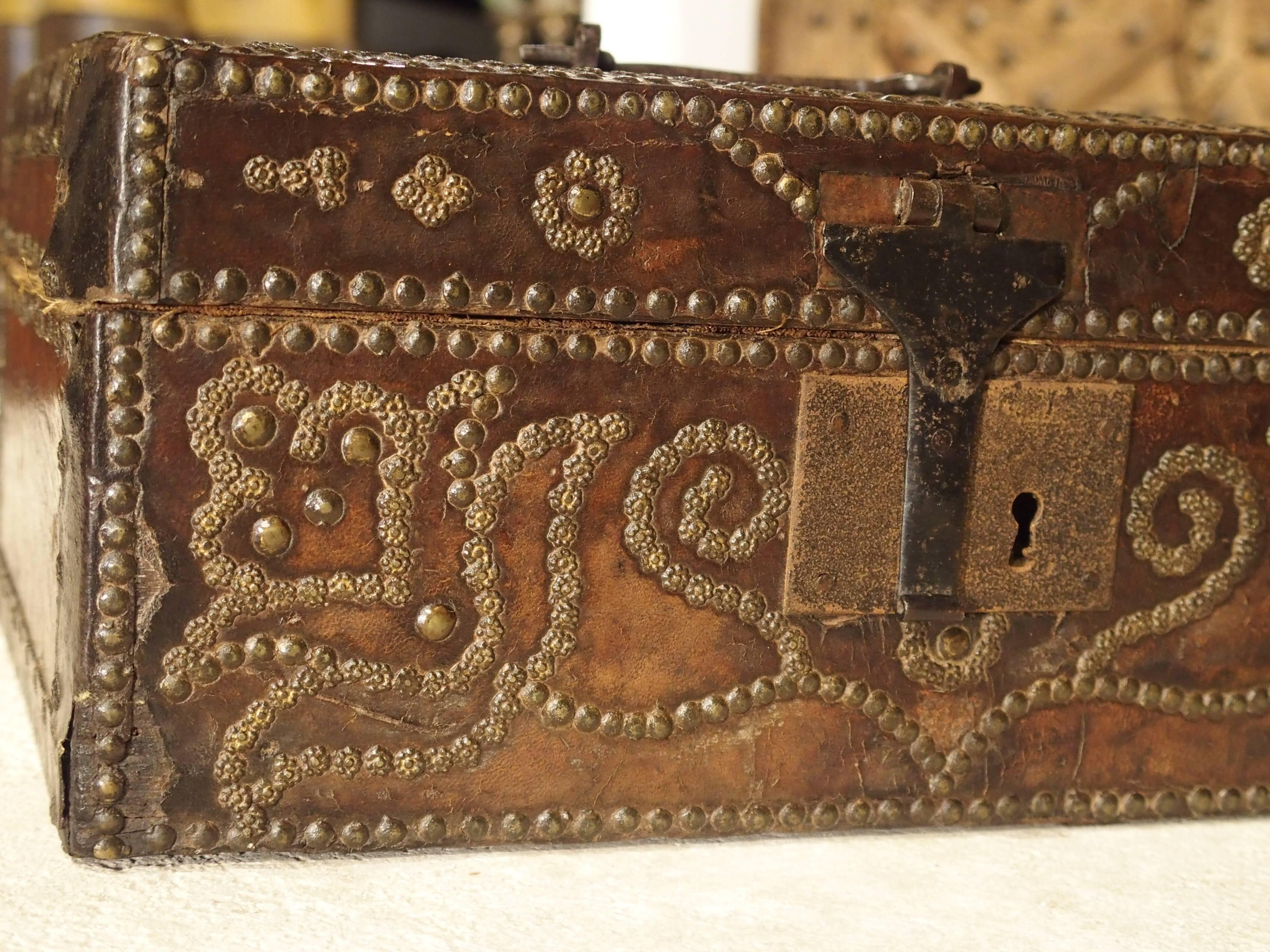 This small 17th century brass studded leather box is from France. There are two types of brass studs used on it, decorative and round. Both are mostly seen on the front making up intricate patterns of floral and foliate designs. The leather is very