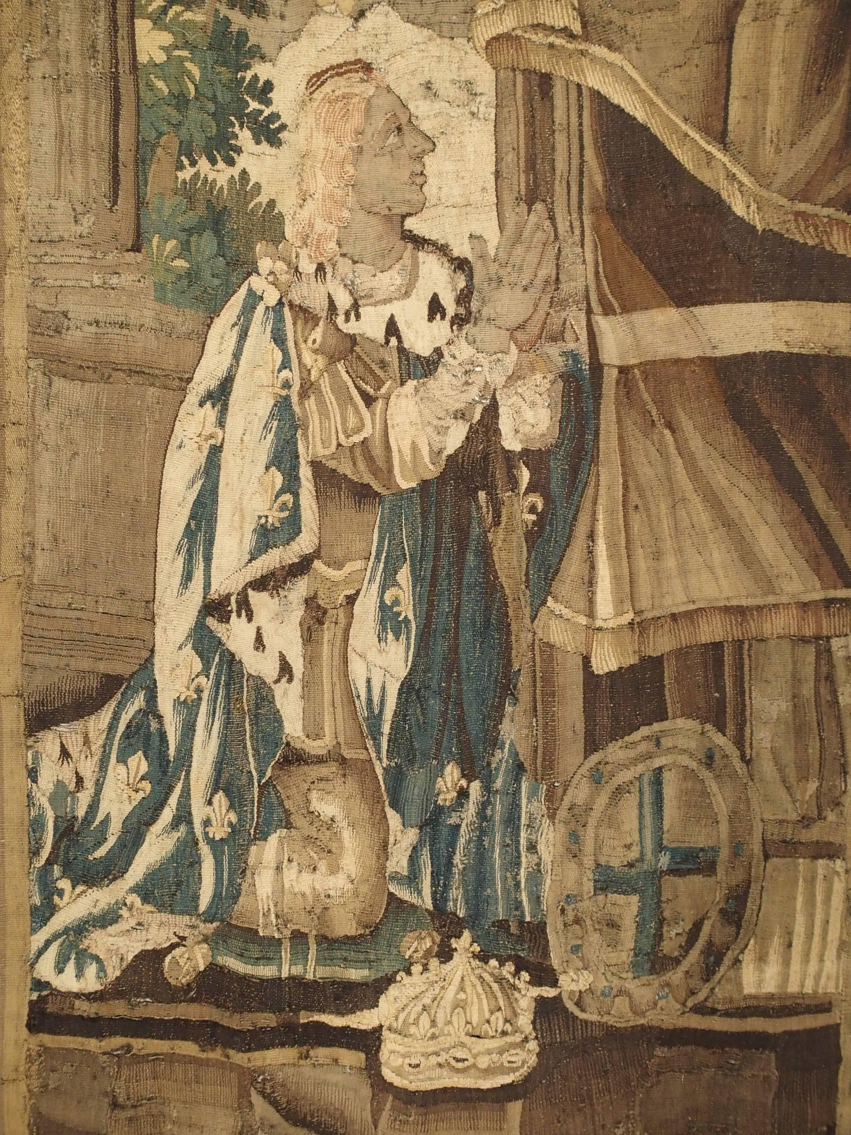 This Aubusson tapestry depicts the coronation a King, kneeling before a section of layered drapery. He is dressed in a Royal robe decorated with the fleur-de-lis motif. His crown is to the right of his knee and it too, has a fleur-de-lis on top of