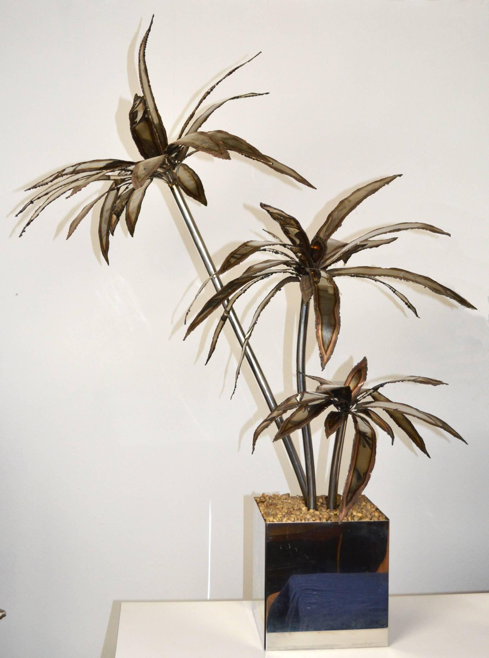An exceptional pair of large steel palm tree sculptures in the manner of Maison Jansen. Three metal trunks with patinated fronds in a polished steel planter with fixed gravel 'mulch.'