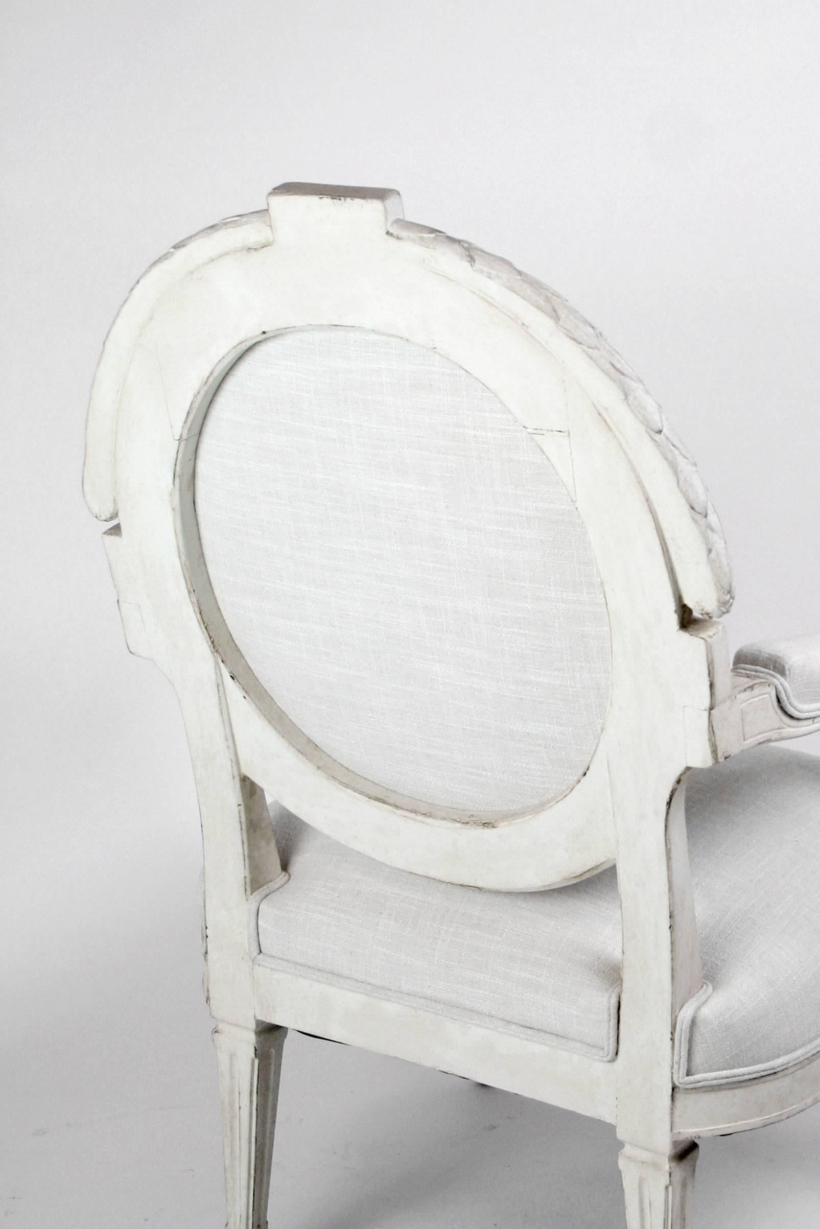 A large and imposing late 1800s, early 1900s French Louis XVI-style armchair with carved wood frame painted in a distressed off-white color. The oval seat back features a carved twisted ribbon surmounted by garland decorations. The legs features