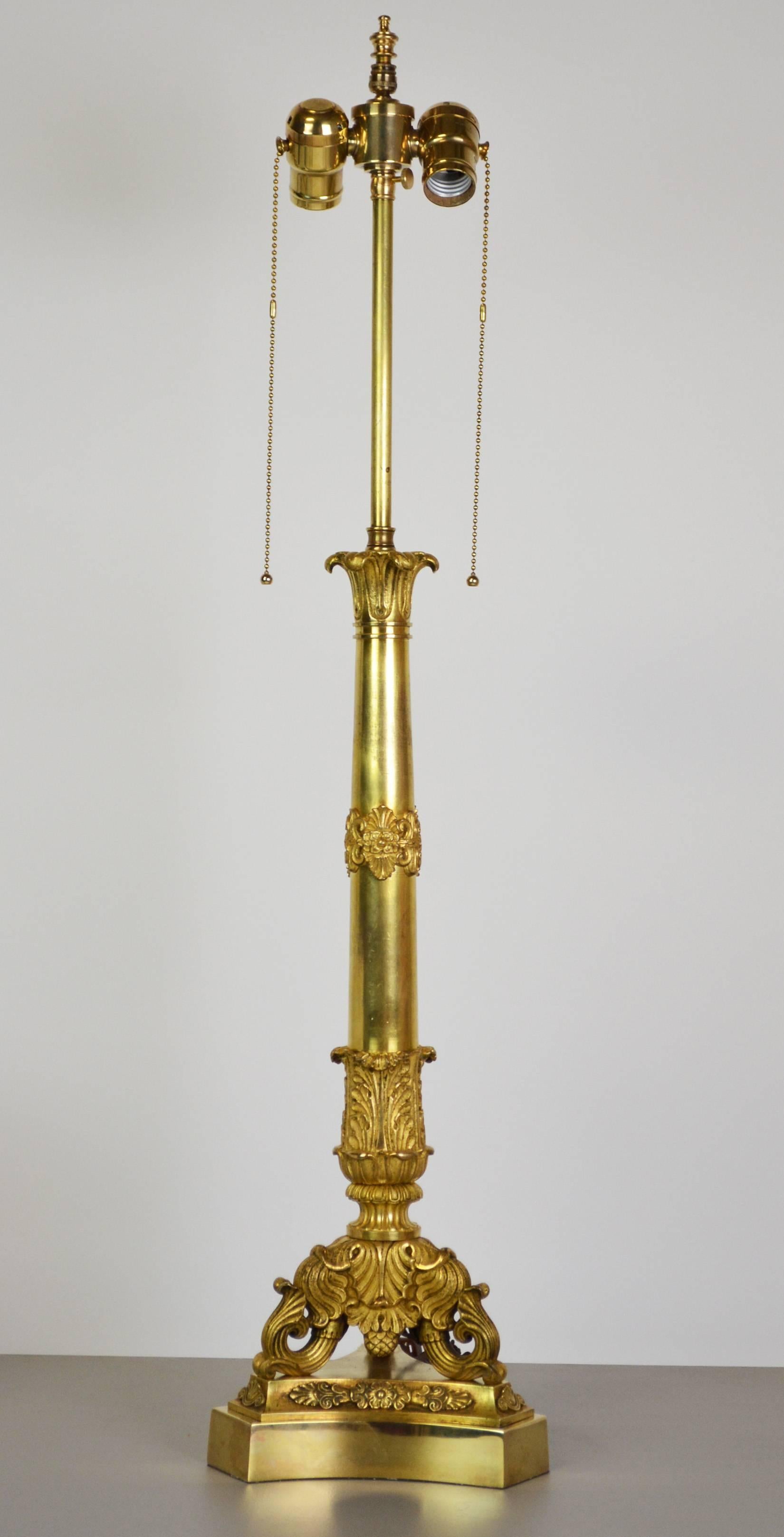 Eine vergoldete Bronzelampe von hoher Qualität aus dem 19. Jahrhundert mit fein ziselierten neoklassizistischen Details. Dreibeinige Basis stützt einen konischen Schaft mit dekorativen Beschlägen, der von einer blütenförmigen Spitze gekrönt wird.