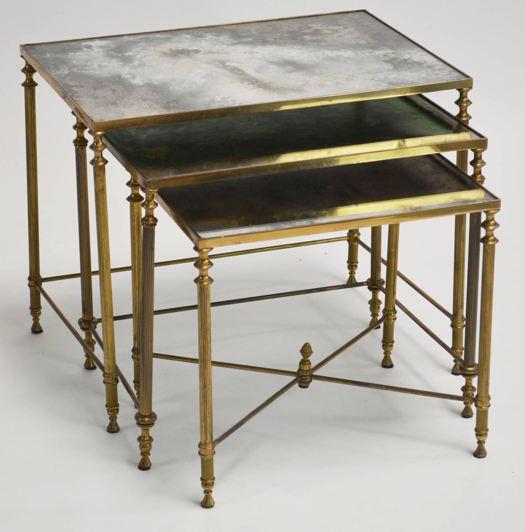 A pair of brass neoclassical nesting tables with original antiqued mirror tops. Typical of French design, possibly Italian, in the style of Baguès and Maison Jansen. Lovely detail, with reeded legs and cast brass finials.