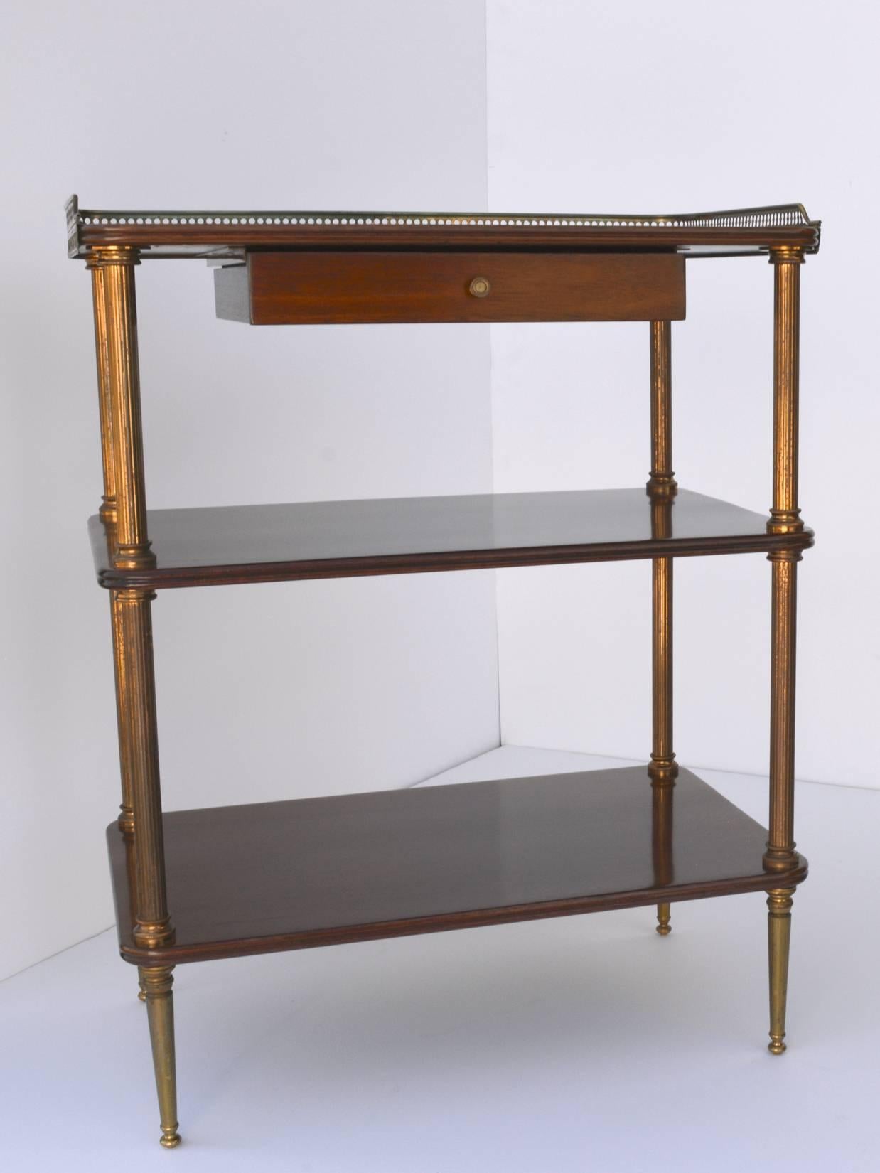 A fine pair of Mid-Century French neoclassical side tables, with three mahogany shelves supported on fluted brass columns with decorative capitals, resting on tapered brass legs. The top has a three sided pierced brass gallery and a small drawer
