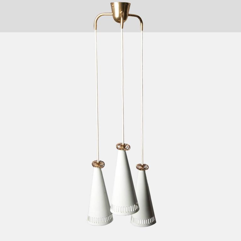 A three-pendant chandelier of brass and white enamel. Shades have a slotted metal detail and are topped with a curled brass ring. By Mauri Almari for Idman, and produced in the 1960s in Finland.
 
  