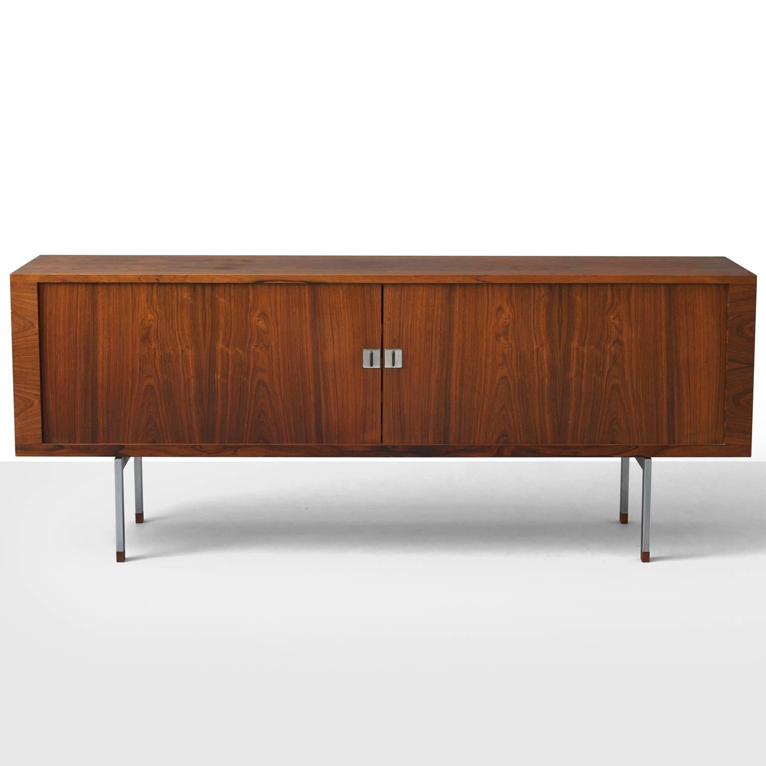 A low sideboard by H. J. Wegner. Features a Brazilian rosewood front with two tambour doors enclosing oak shelves and sliding trays. Chromed metal base with rosewood leg ends. Produced by Ry Møbler. Includes certificate from the Danish forest and