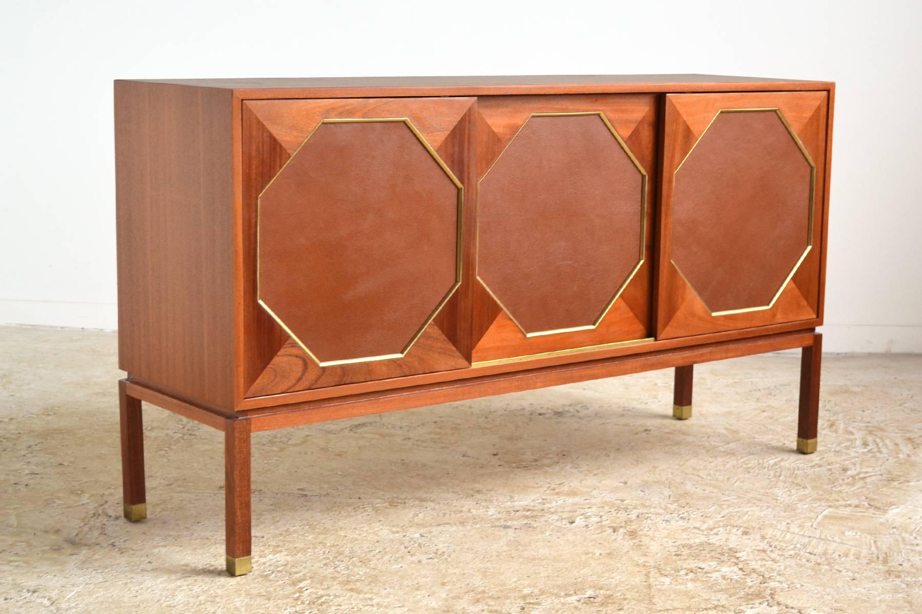 Harvey Probber's refined aesthetic shines in this three-door cabinet. Crafted of mahogany, it has three sliding doors with octagonal leather inserts framed in brass which conceal two compartments with two shallow slide-out drawers/ shelves and one