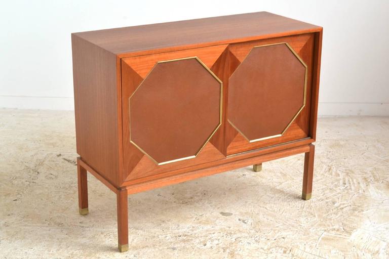 Harvey Probber's refined aesthetic is on display in this three-door cabinet. Crafted of mahogany, it has two sliding doors with octagonal leather inserts framed in brass which conceal two compartments, one with two shallow slide-out drawers/ shelves
