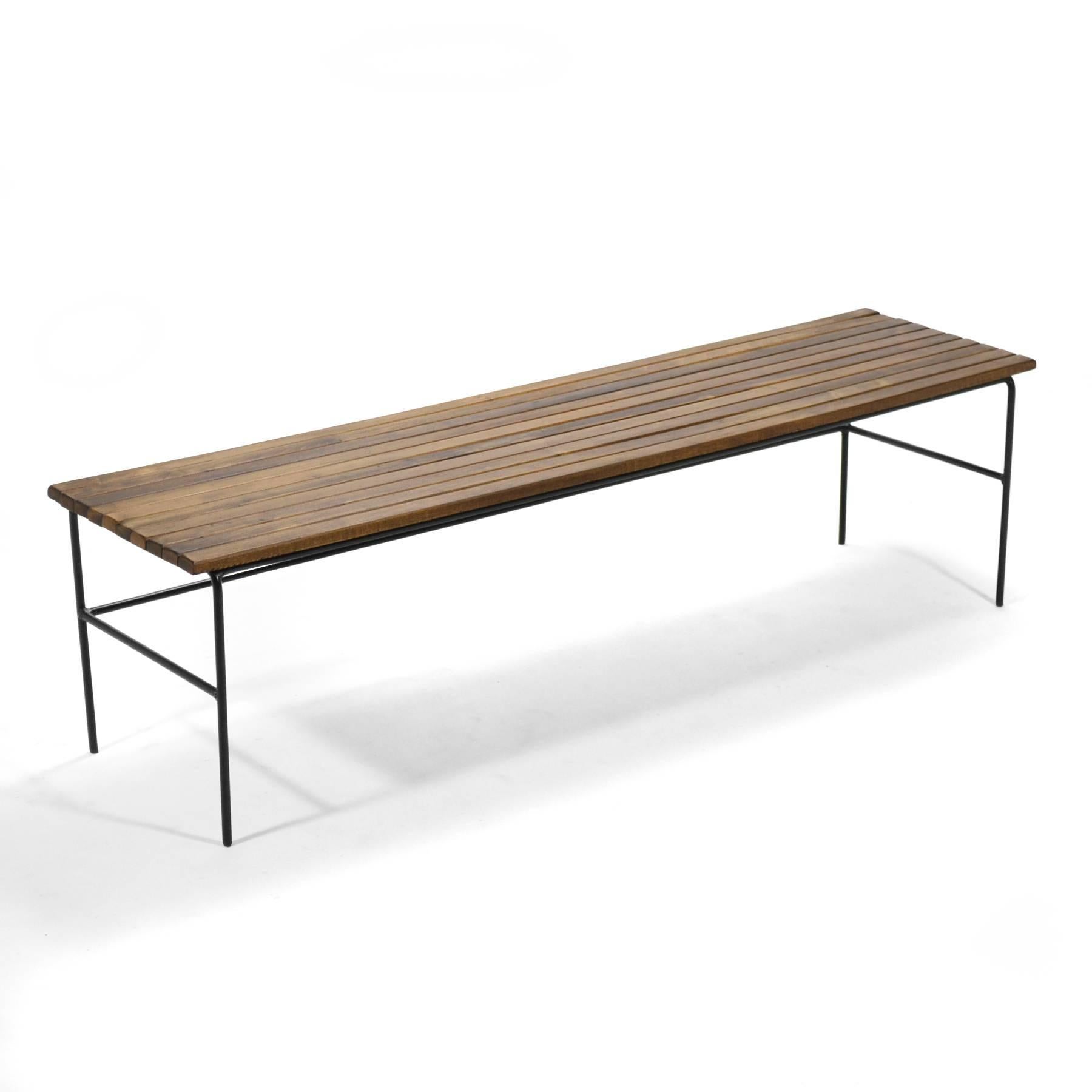 The slat bench was a very popular form in Mid-Century America. Highly functional, it can serve as a seat or a table. This design by Arthur Umanoff has a top of wooden slats supported by a simple iron base. The piece has a strong graphic quality with