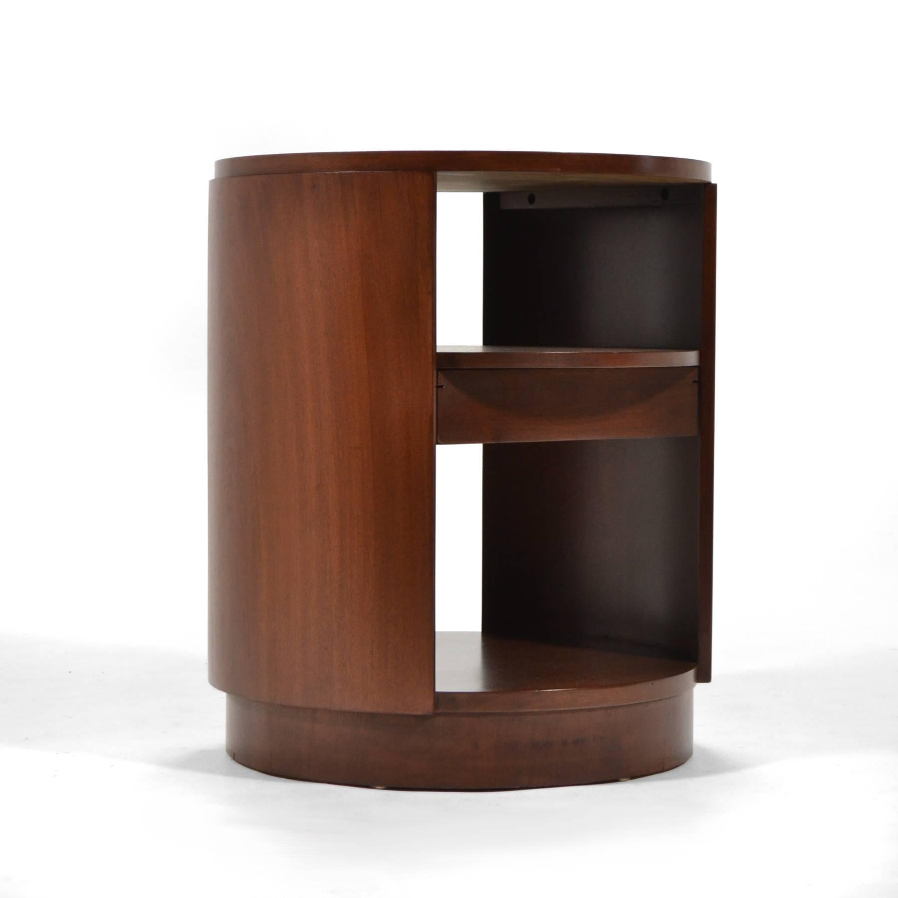 A beautiful design by Ed Wormley that references deco and modern aesthetics, this rare side table has a cylindrical form and a drawer below the middle shelf. Expertly crafted of mahogany by Dunbar and newly restored.