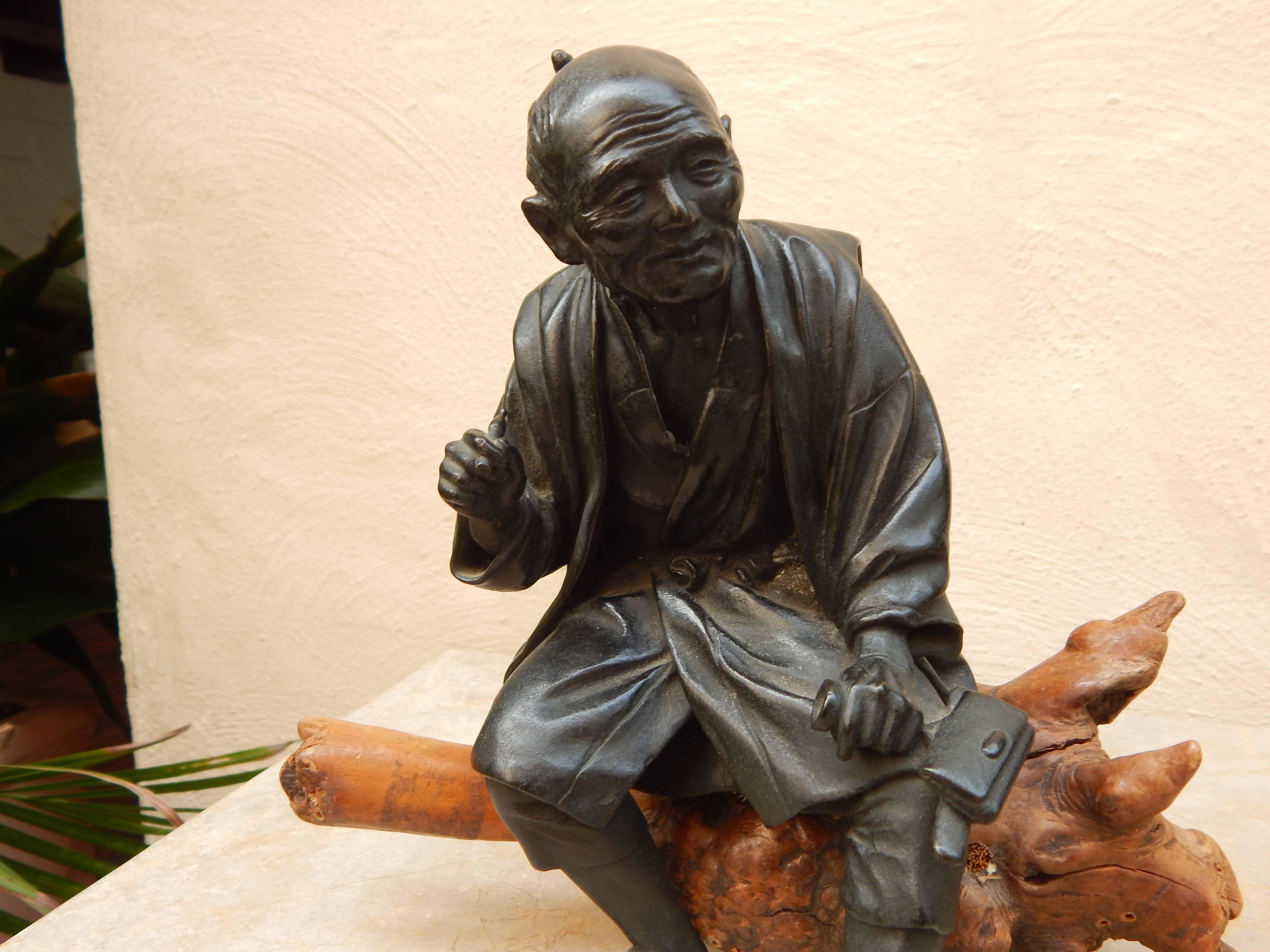 Very unique black patinated bronze figure of a resting Asian elder man on separate weathered burl wood base. Figure is dressed in period attire, with elevated hand holding a pipe and with folded purse in the other.