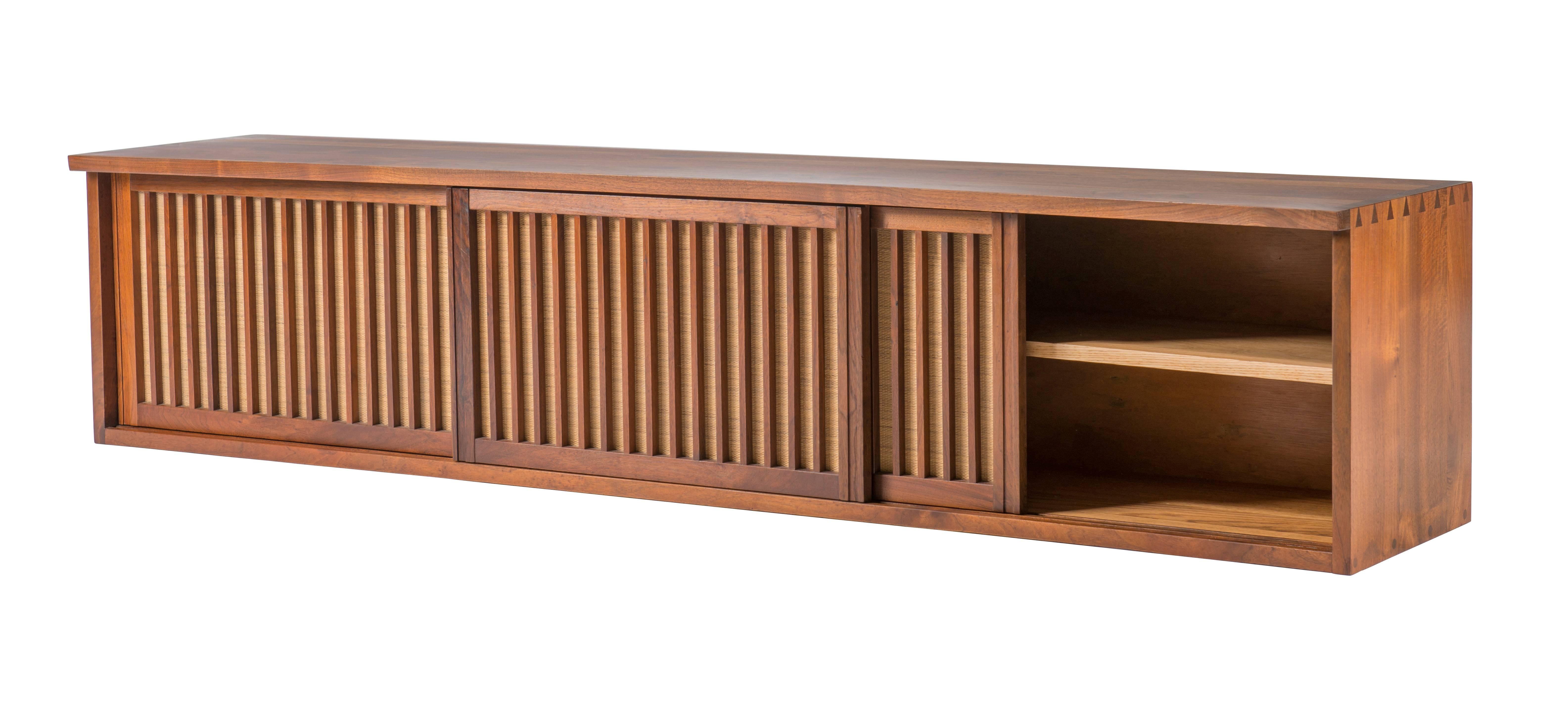Beautiful Nakashima 7 Ft. wall-hanging cabinet in walnut with a free-edge top and exposed joinery at cabinets edges. Cabinet with three sliding pandanus cloth covered doors revealing two shelves and four small drawers. Signed with original owners