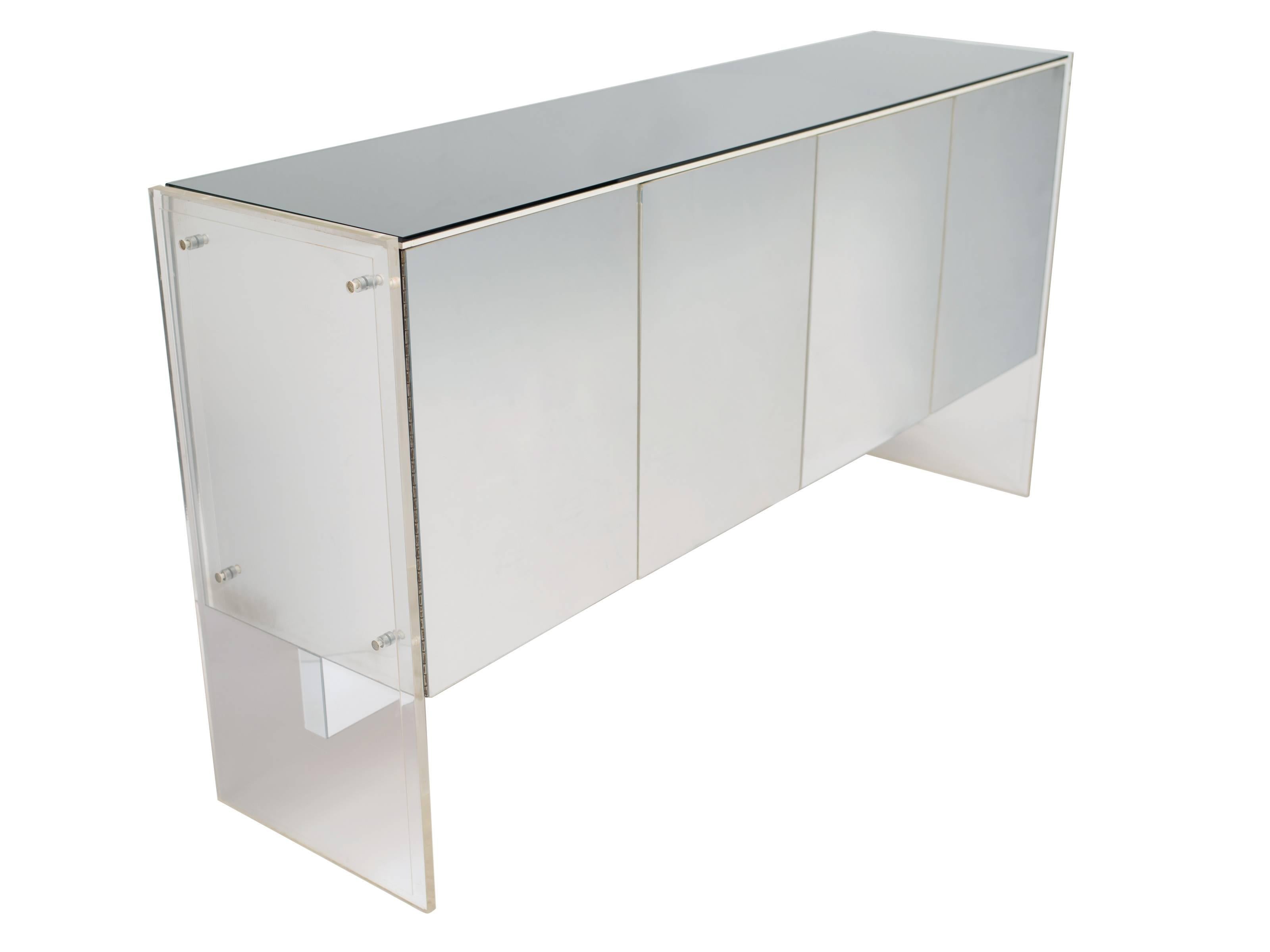 Modernist sideboard in Lucite and glass. The credenza is comprised of four reflective metal door fronts and thick Lucite sides/frame with stylized bolts. Fitted with a beautiful smoked mirrored top. The doors open to reveal interior cabinet and