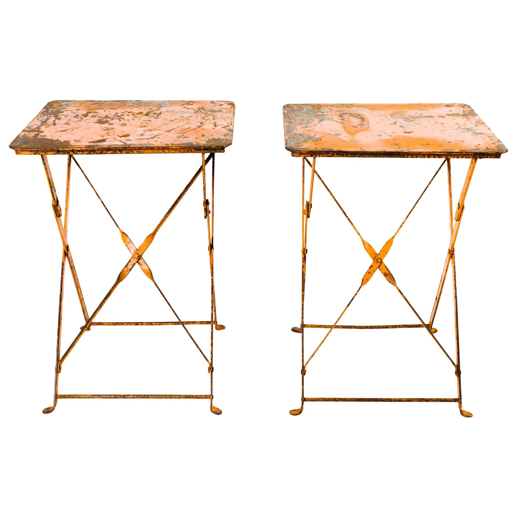 Pair of industrial French folding iron bistro tables with beautiful patina on the metal and original orange enamel paint. Exhibits wear and distress on the paint throughout, adding to their rustic charm and beauty. Can be used indoors as side tables