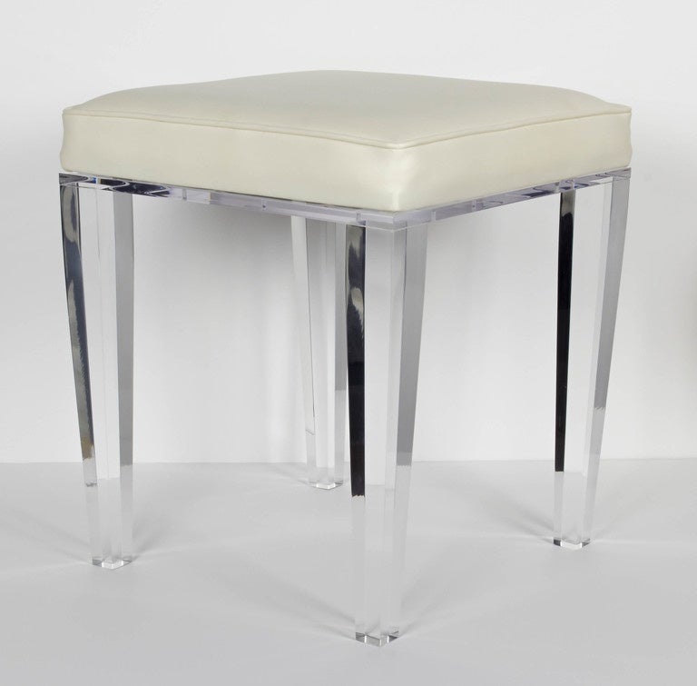 Elegant and modern lucite vanity stool with luxury leather seat in crème. The stool has a square form with a beautiful Lucite base comprised of four tapered legs with beveled details. Perfect scale for a vanity, a powder room, or dressing room.