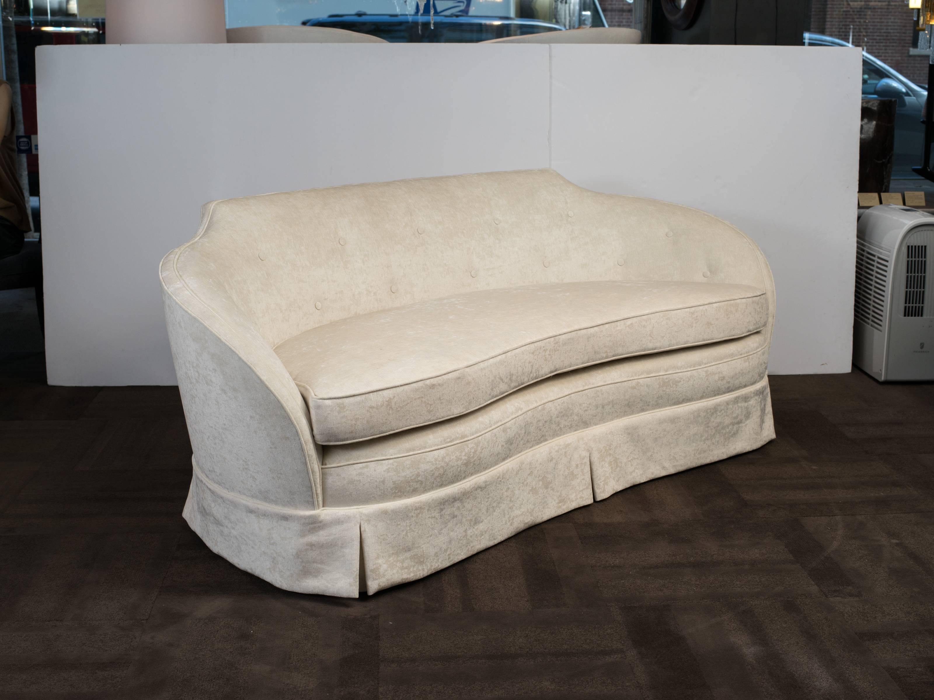 Striking Hollywood era sofa with stylized curved back/arm design. Newly reupholstered in a rich crème velvet by Castel, featuring button back details and dust ruffle. Comfortably sits up to four people.