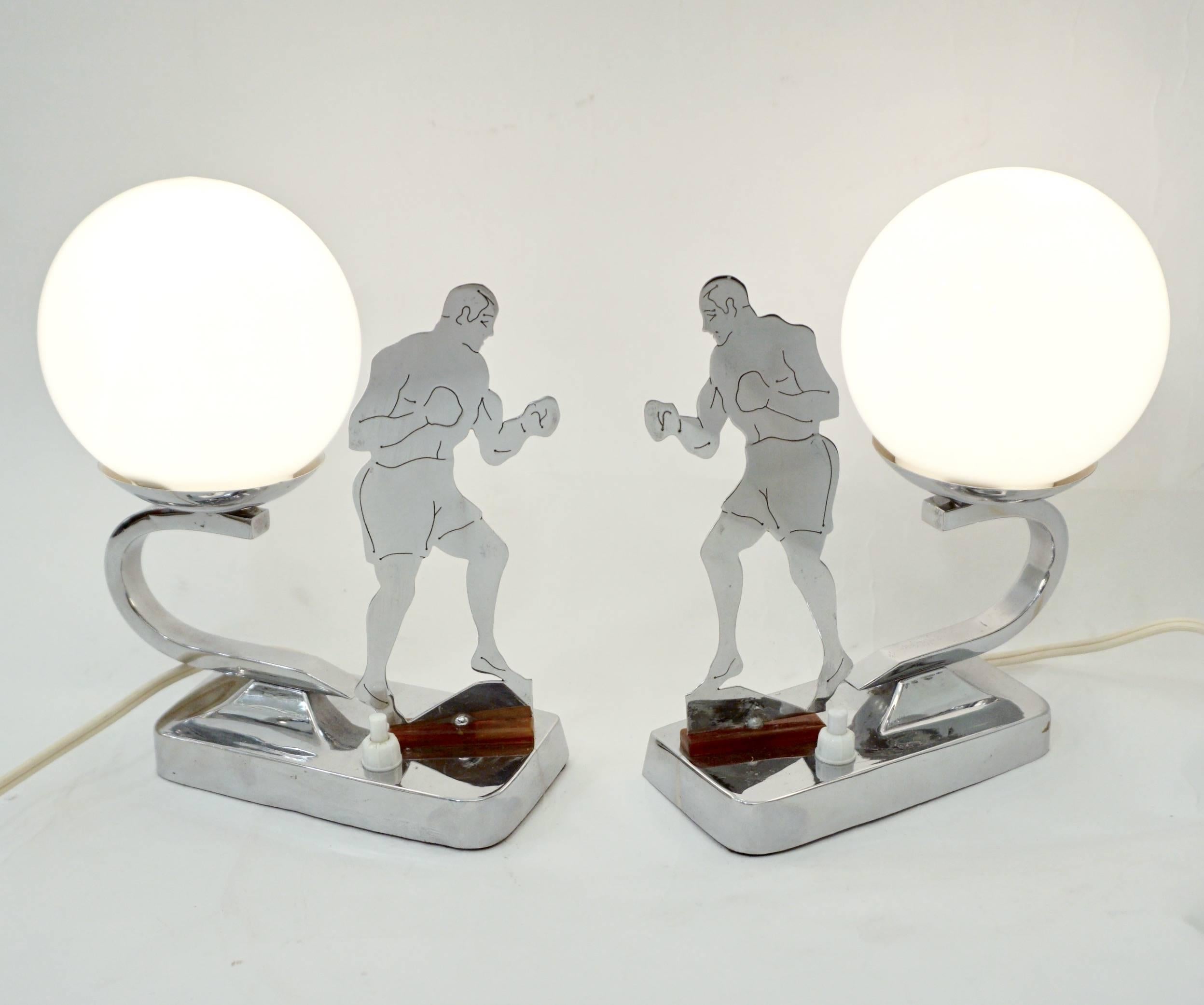 A charming pair of silver color nightstand / side table lamps, handmade in Great Britain, representing Minimalist boxing opponents, chrome cut-out figure sculptures raised on a small bakelite base, the blown glass globes supported by a curved