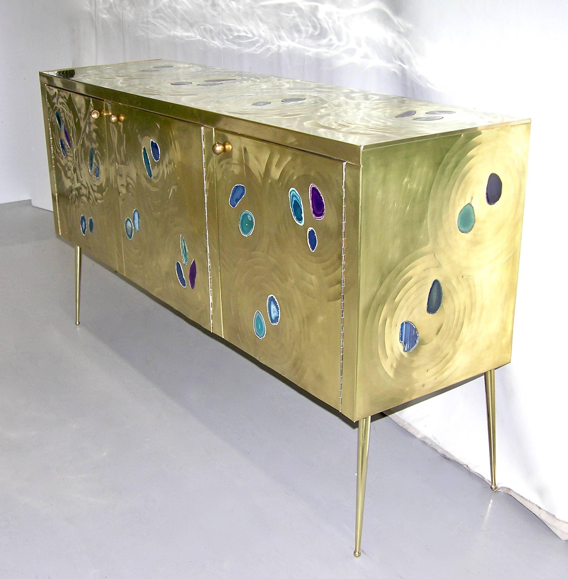 A fine design Italian credenza or sideboard with three doors, entirely handmade, wrapped in gold brass decorated with insets of cut agate in purple, blue and green. The stone is set like jewelry in the brass with a sophisticated rippled decor like