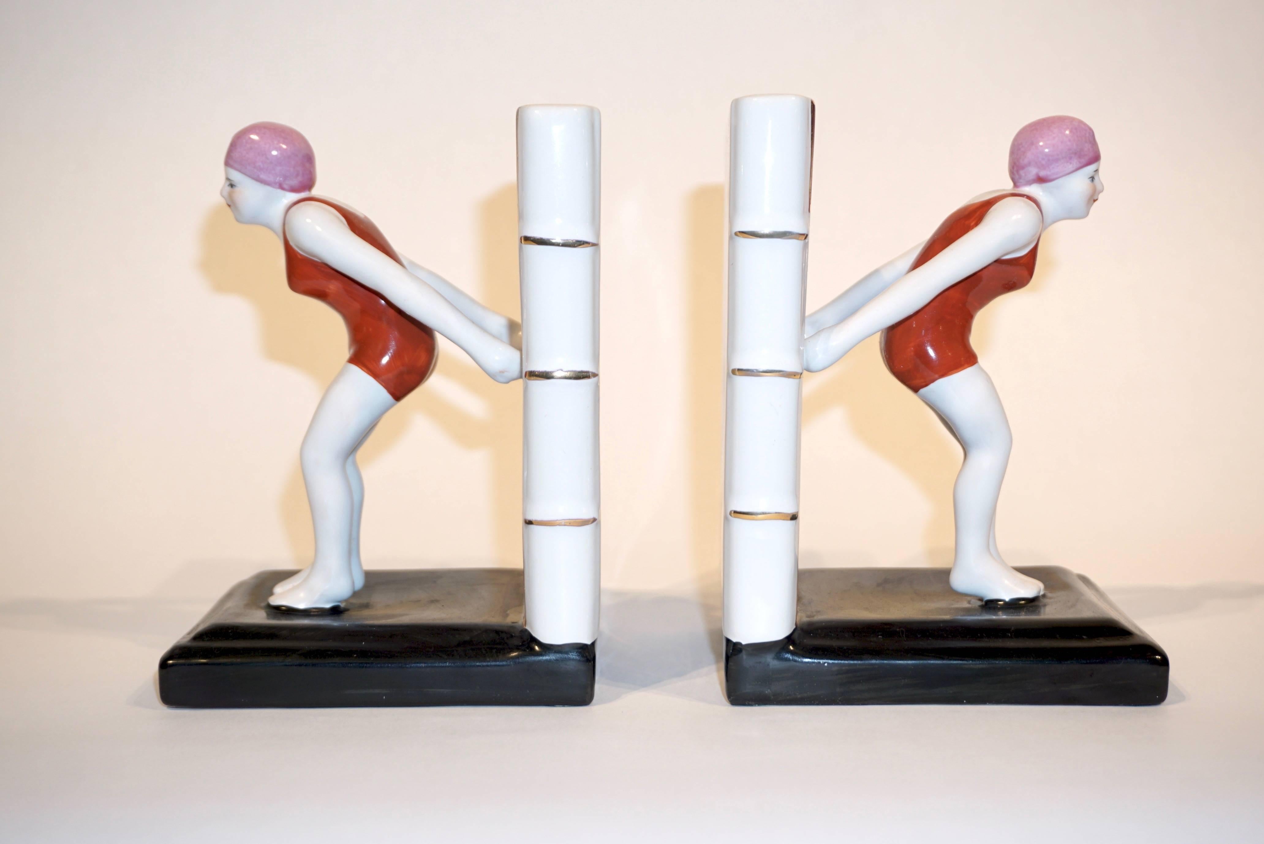 A delightful pair of ceramic Art Deco bookends representing each a diving female sculpture in red bathing suit and pink swimming cap raised on a black plinth, supported by a white back representing a book with yellow edges.