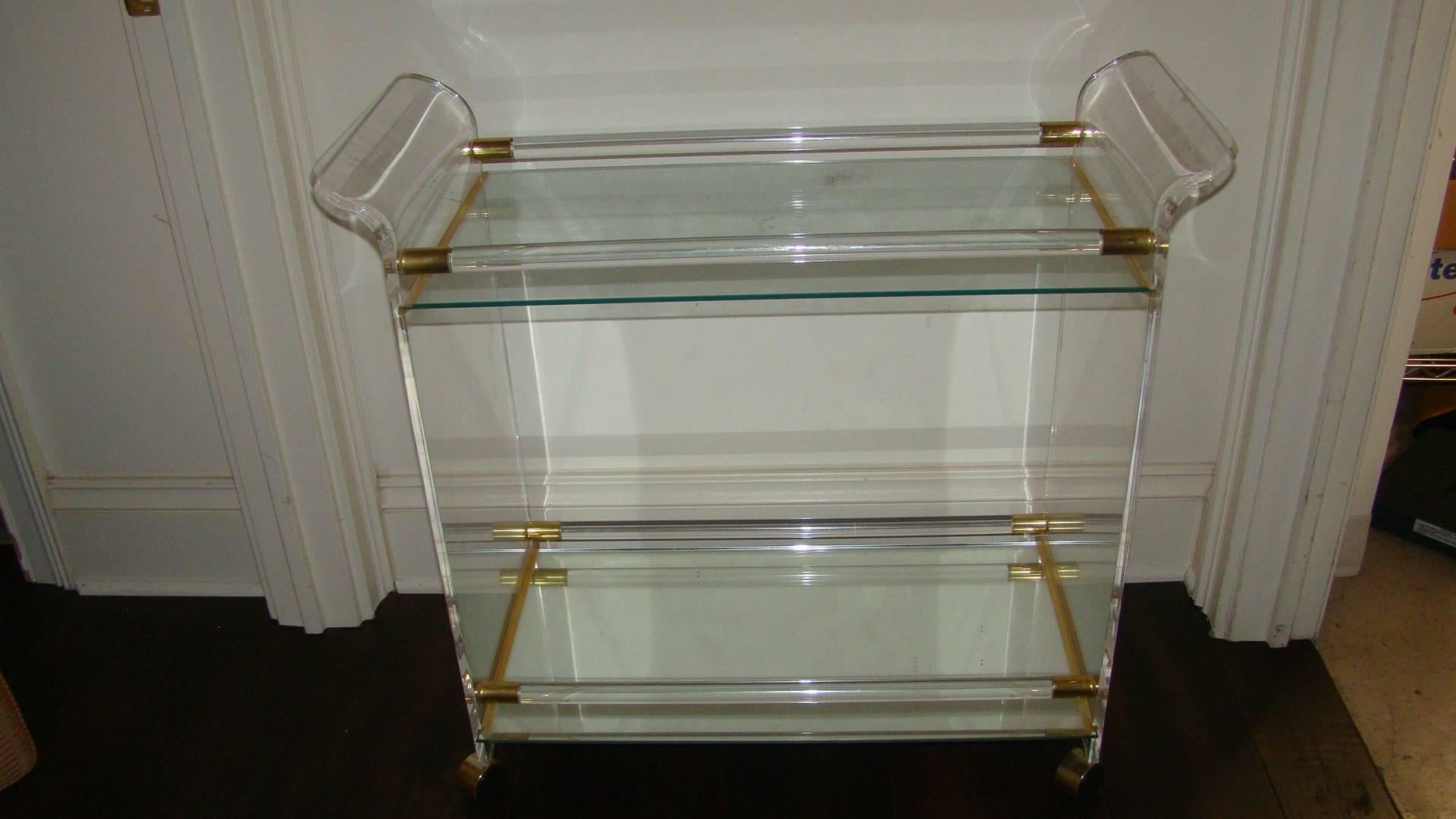 Beautiful Lucite and brass sculptural bar cart. Comprised of crystal clear Lucite with waterfall style designed handles and brass accents. Bottom shelf is mirror, top is glass. Perfect for bar or serving.