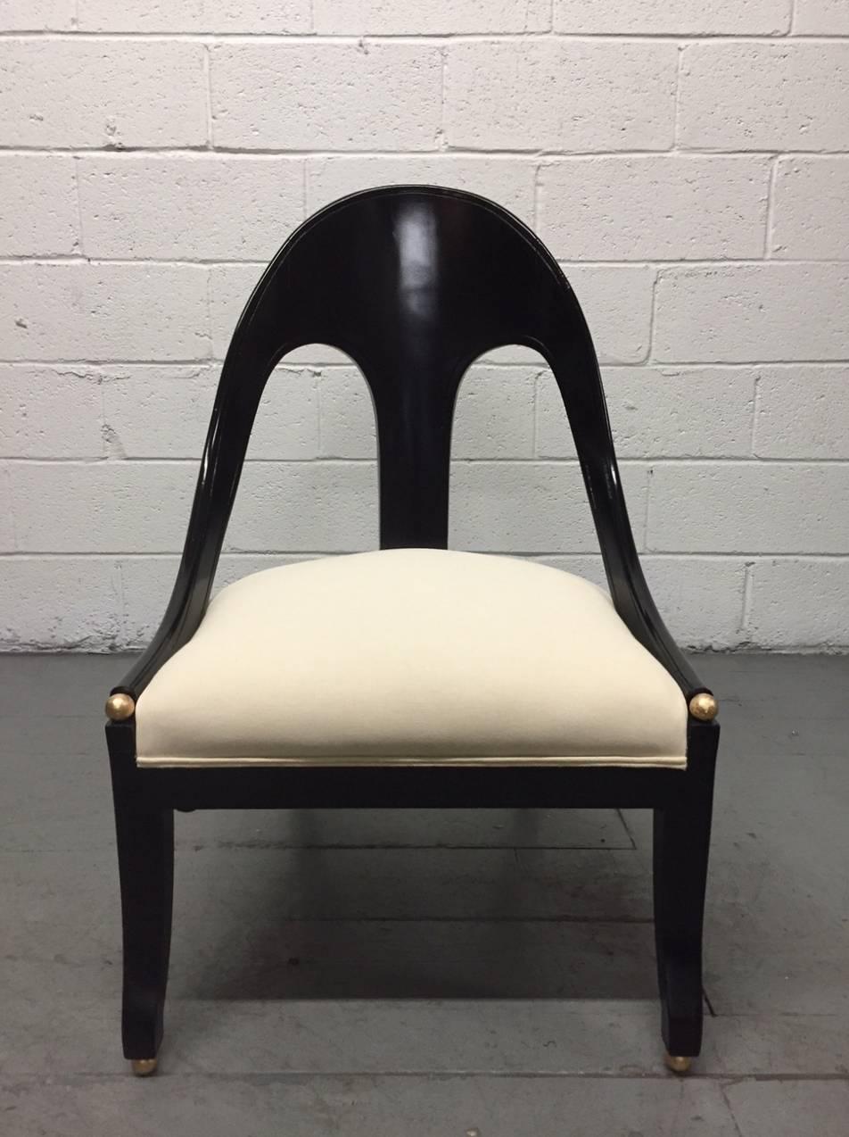 Pair of neoclassical style spoon back lounge or slipper chairs. Chairs have a black lacquered finish. To the front of the arms and feet, are gold ball finials. Seats newly reupholstered in a linen-blend fabric. 