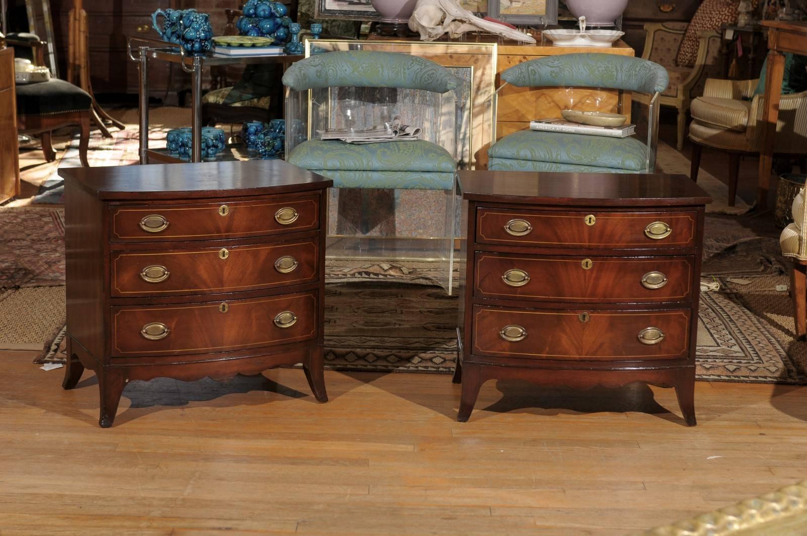 Pair of early 20th century American petite bow front chests in the Georgian style and made of flame mahogany having three graduated drawers with satinwood inlay and brass escutcheons and bail hardware.