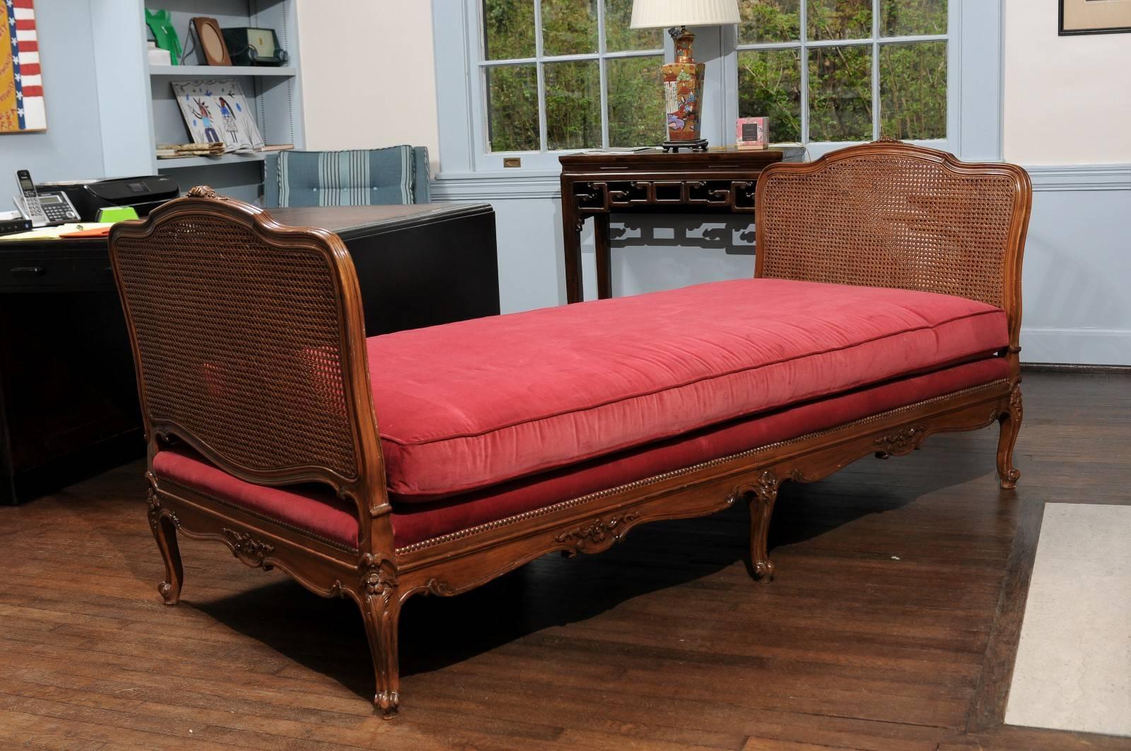 19th Century French Provençal daybed of cane and walnut in the style of Louis XV. The custom red velvet cushion rests on a platform with six carved cabriole legs joined buy floral carved and scalloped aprons and bordered by decorative nailheads. The