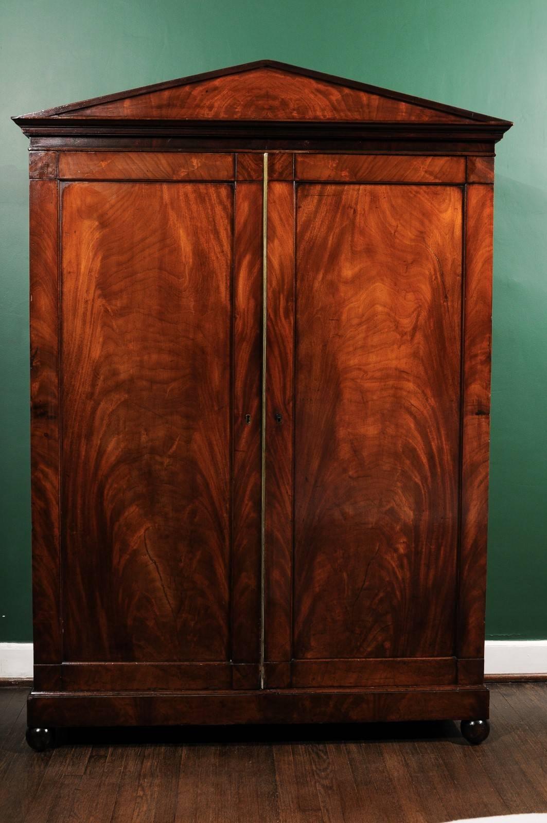 19th century French armoire of the Louis Philippe period covered in a flame mahogany veneer. The armoire has a triangular crown over two large doors with brass hardware that open to a fitted case. On the left are the original mahogany veneered