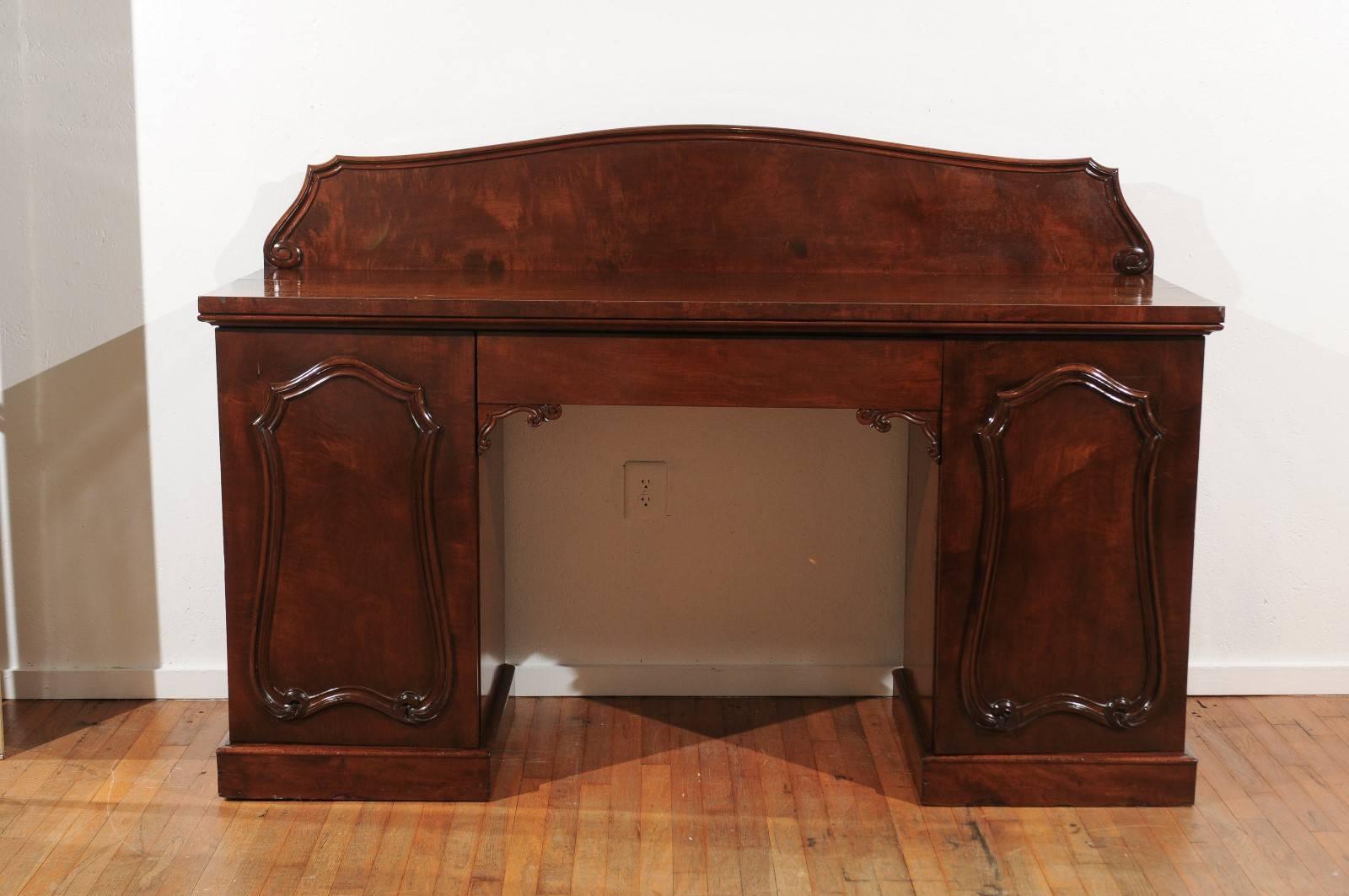 19th century sideboard of mahogany in the style of William IV. The sideboard has a thick mahogany top holding a wide drawer in its frieze, and is supported by two pedestals standing on plinth bases. Each pedestal has a door with carved moldings and