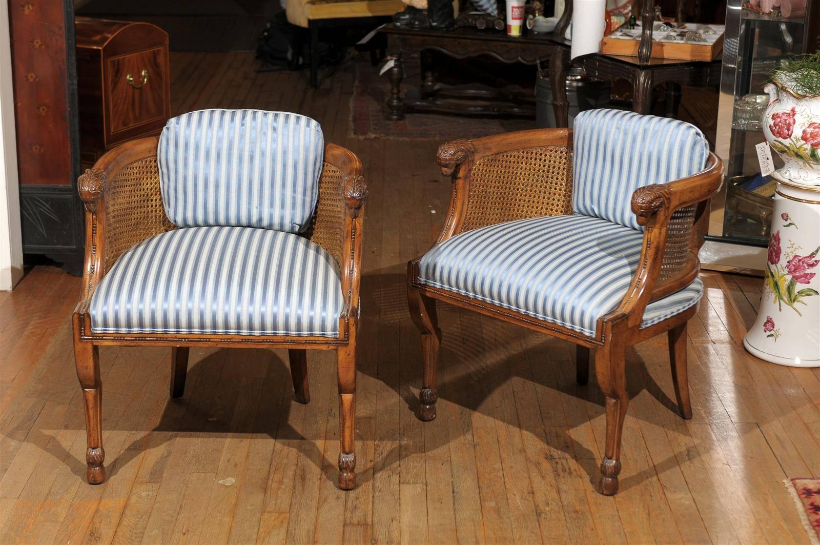 Pair of Mid-Century Regency style club chairs of solid walnut with carved ram's heads at the arms' handholds, cloven front feet and saber style back legs. The barrel back features the original cane. The seats are upholstered in a light and dark blue
