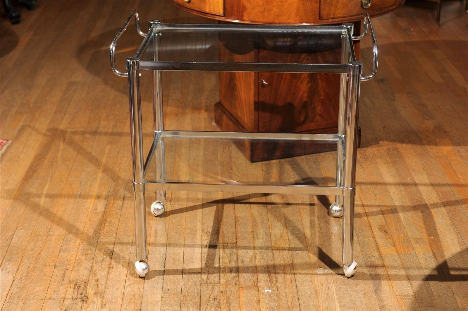 Midcentury bar cart of chrome having two tiers holding glass panels and upturned handles on either side.  The cart is raised on wheels and the upper tier raises off of the lower making it easier to store or ship.  