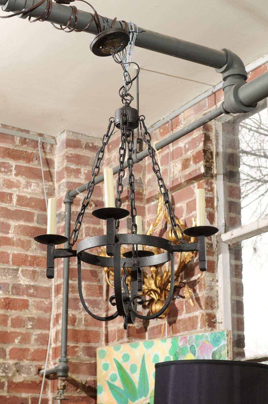 This 20th century chandelier is made of black iron in the Spanish or Mediterranean Mission style. It has a decorative ceiling plate holding a single chain that drops 9 inches to a small pendant. The pendant supports four chains that form a basket
