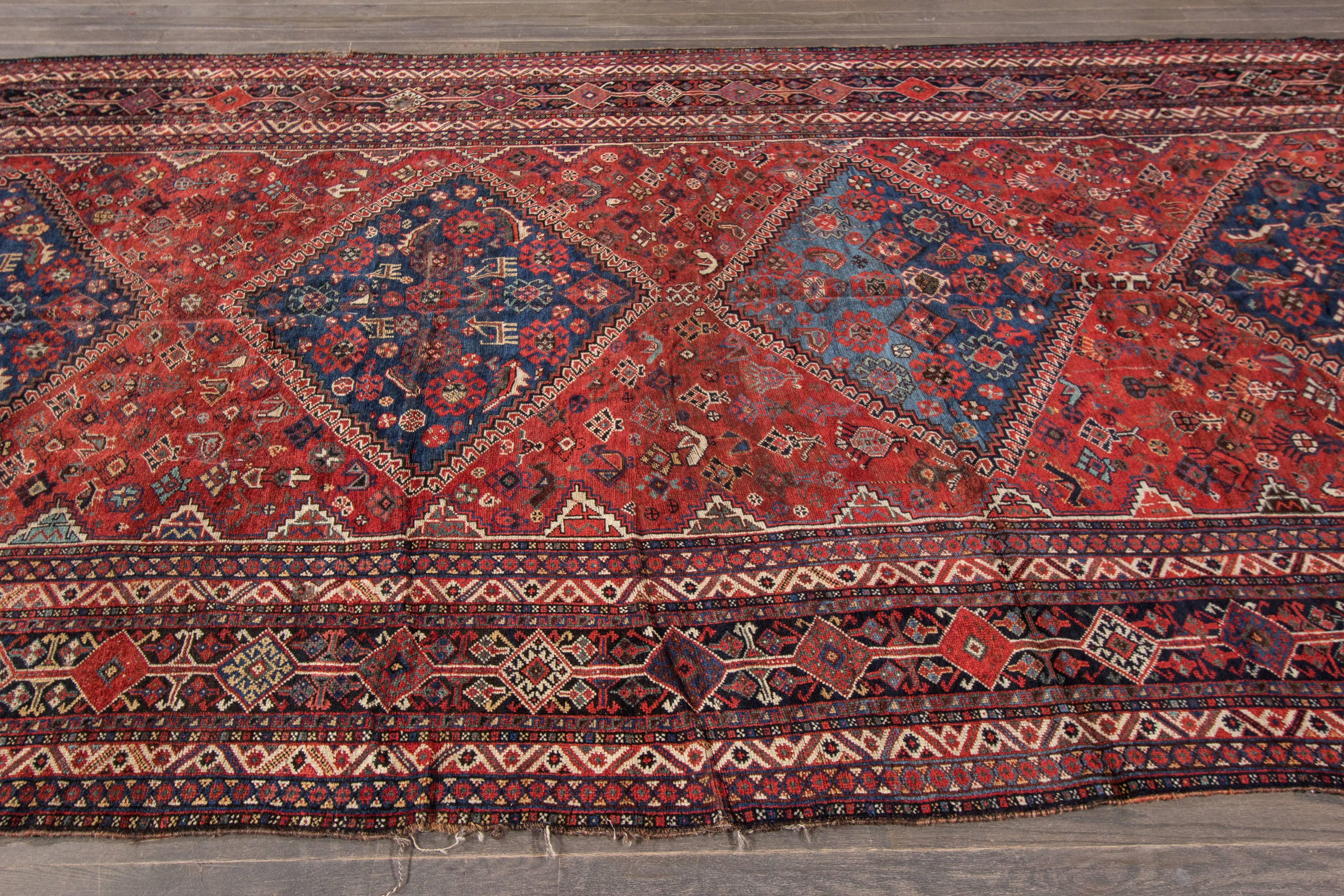 A hand-knotted antique Shiraz rug with a geometric floral pattern on a red field. Accents of ivory, blue and beige throughout the piece. The size of this piece is 6'.11 x 15'.7.