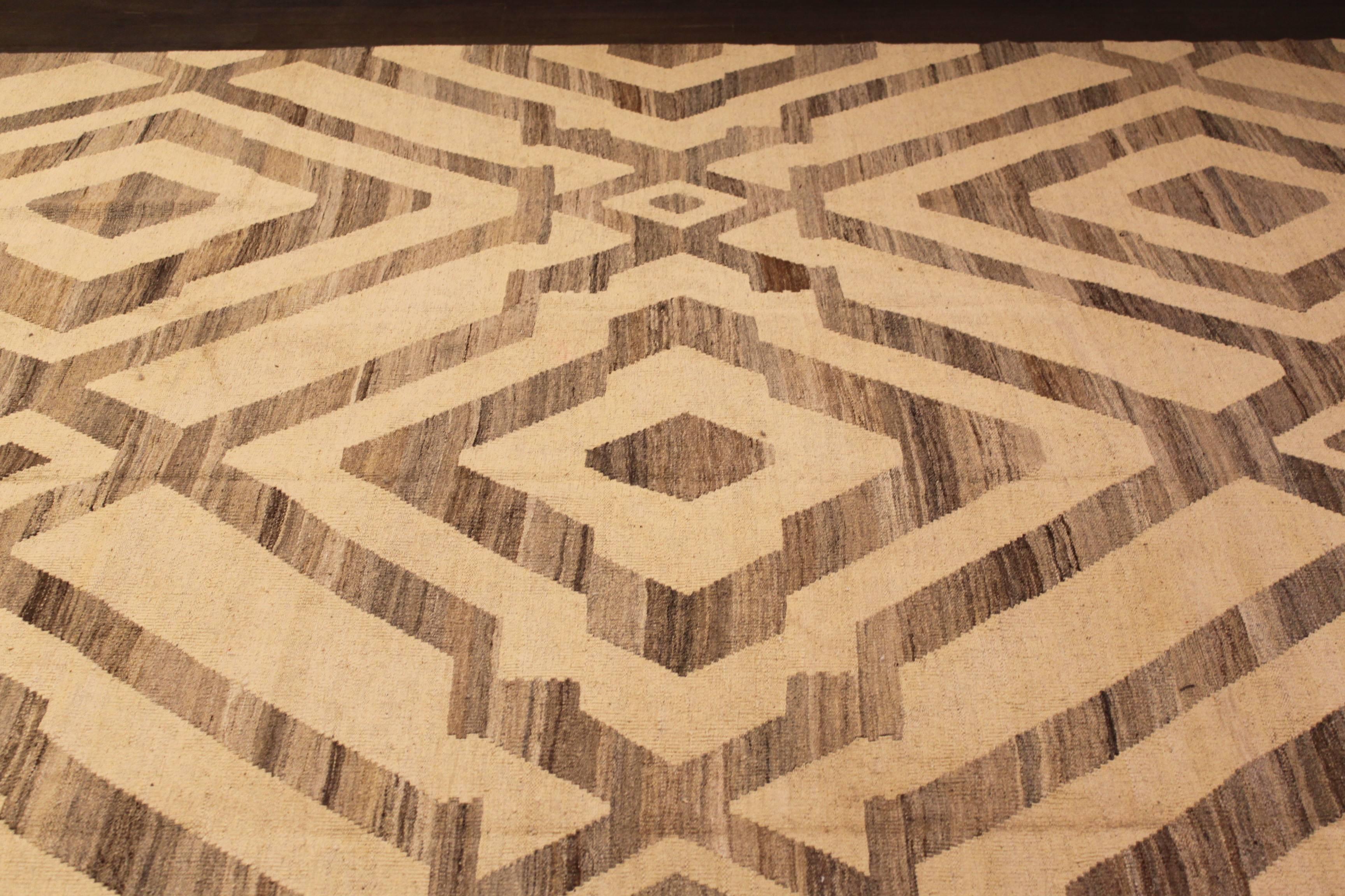 Un tapis Kilim noué à la main avec un design géométrique sur un champ beige. Accents de brun et de beige dans toute la pièce. Ce tapis mesure 9'.2 x 12'.5.
