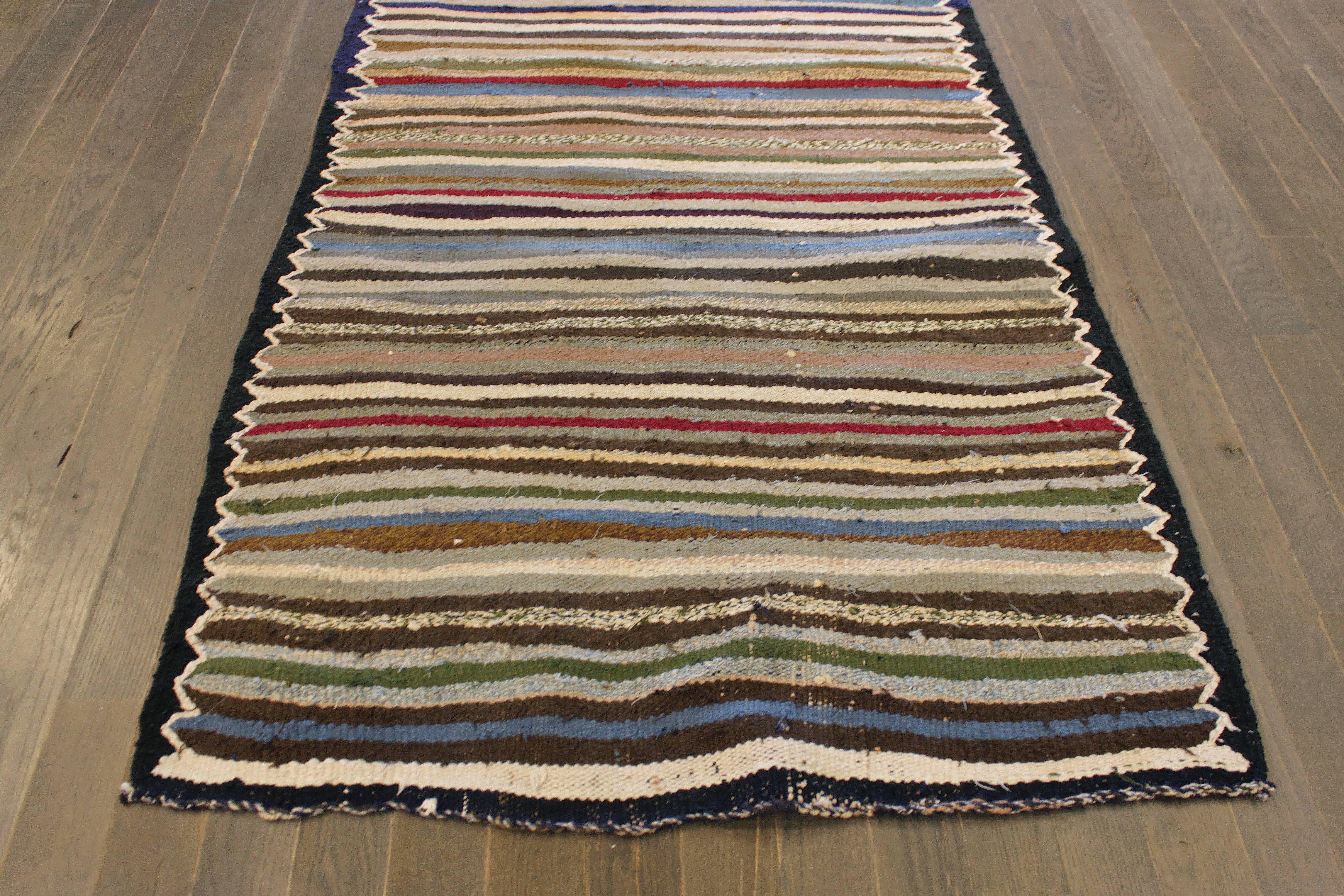 A hand-knotted Kilim rug with a striped design on a black field. Accents of ivory, green, red and blue throughout the piece. This rug measures 3'.5 x 9'.4.