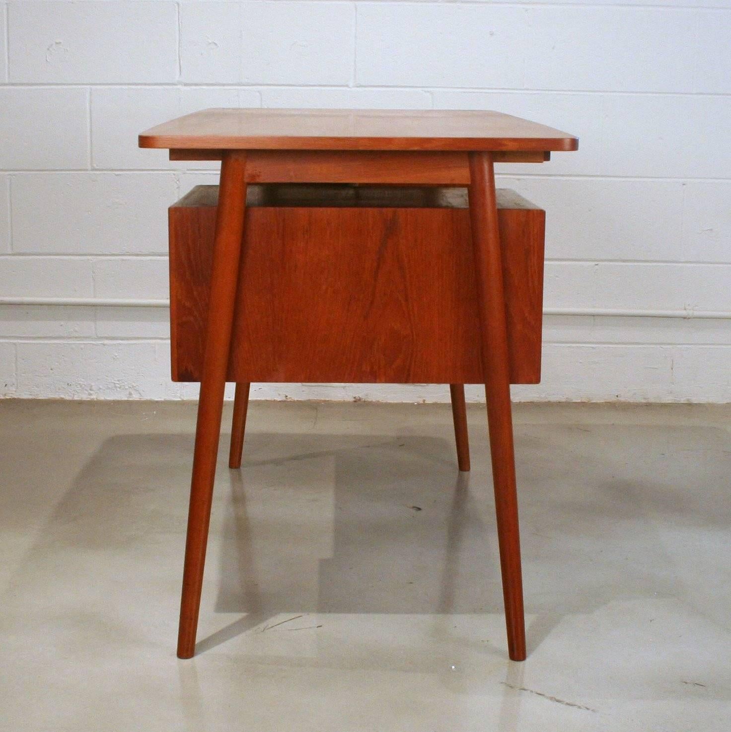 Small-scale vintage Danish teak writing desk with single bank of drawers and small storage shelf. Drawers feature solid teak half-moon pulls. Rear has an open storage shelf. Resting on solid conical angled legs. Made in Denmark.