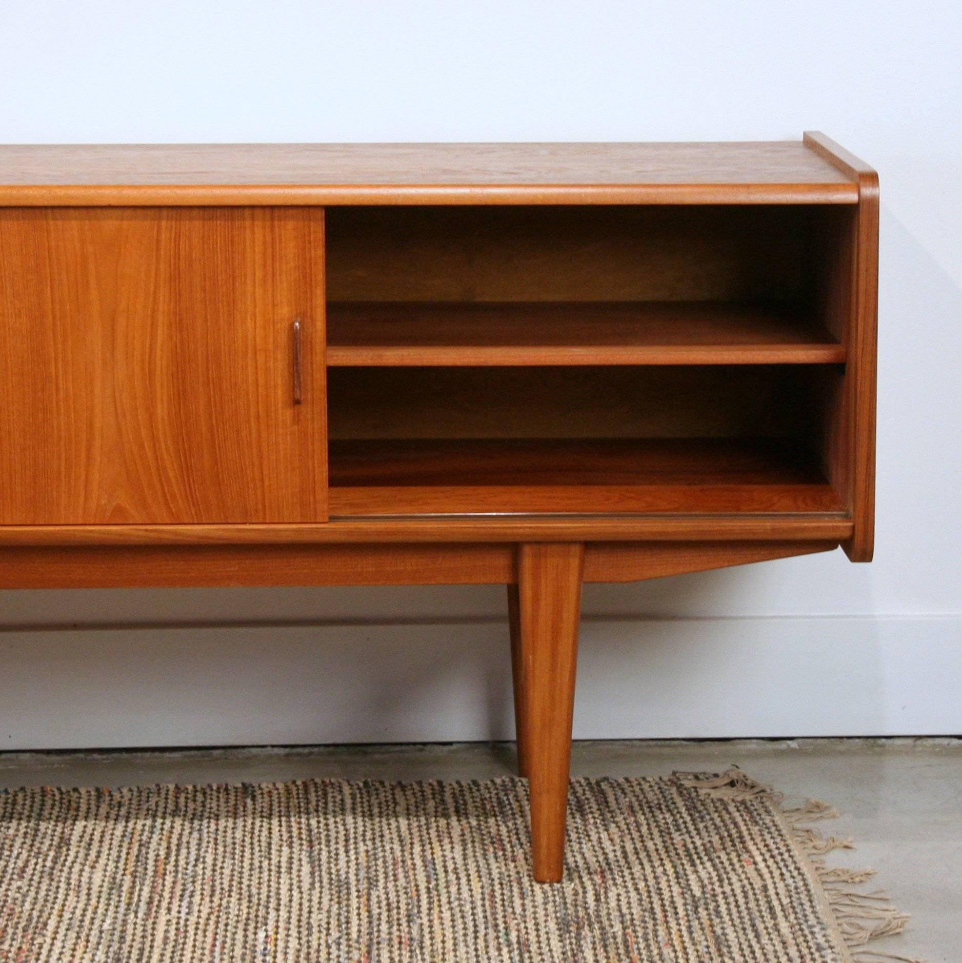 Stunning long vintage Danish teak sideboard with centre bank of drawers. Solid rounded front edging with solid thin, angled teak pulls. Resting on solid tapered teak legs. Sliding doors opening to reveal a removable shelf. Made in Denmark.
     