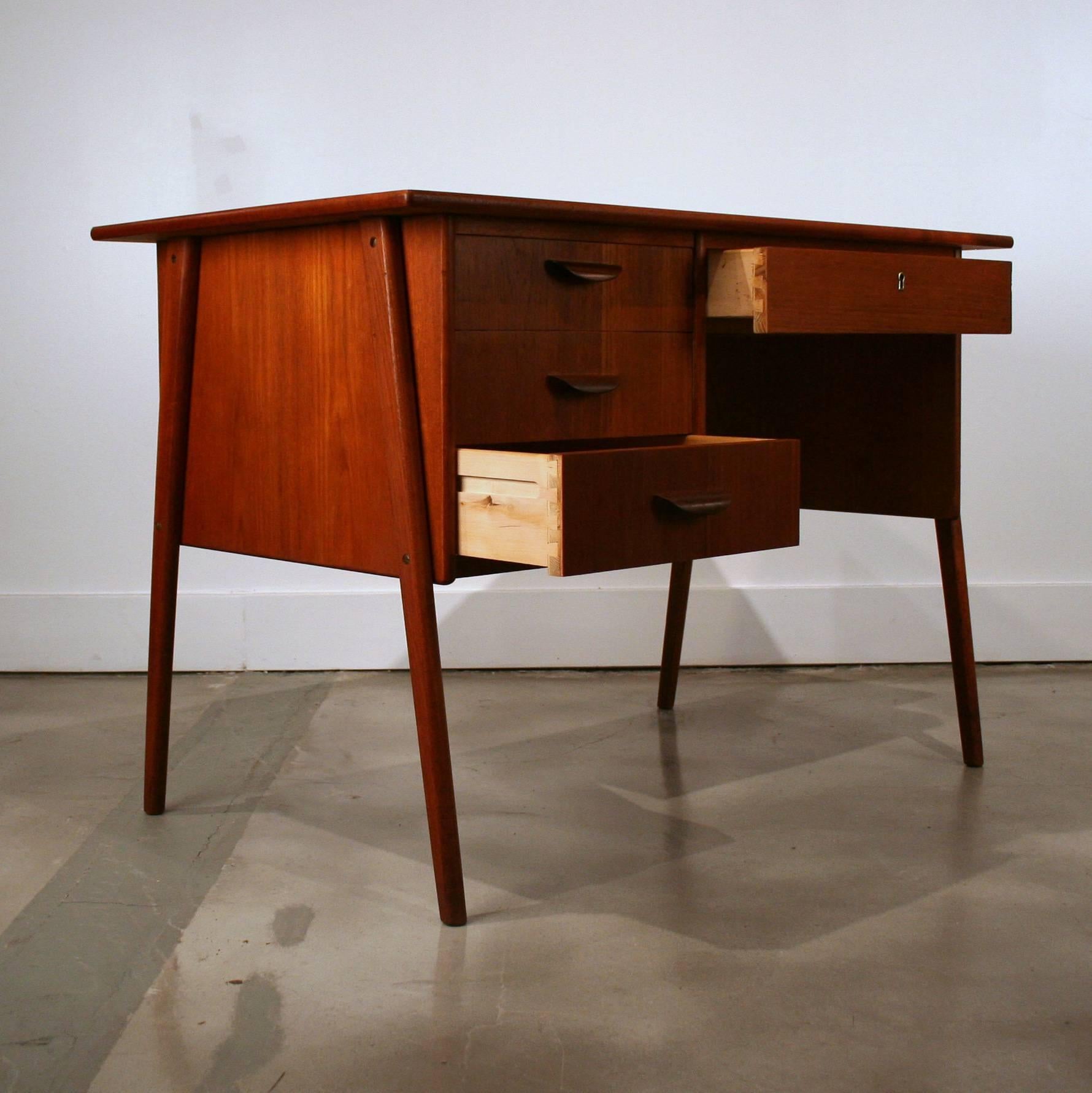 Small vintage Danish teak desk, features three drawers on the left with downward curved finger pulls and one drawer on the right directly below the desk top. This piece also features splayed legs and dovetail joinery. Made in Denmark.