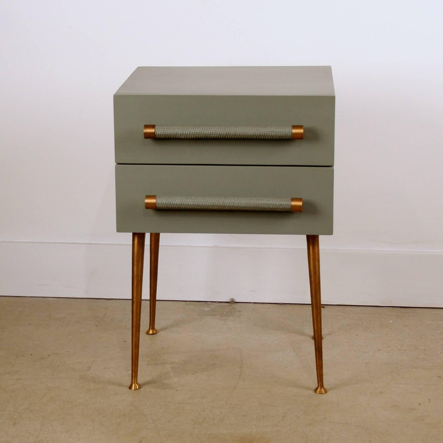 This small two-drawer bedside table features one drawer with a wicker handle, brass trim and is set on four brass legs. Inspiration for this piece comes from the 1950s designer Robsjohn-Gibbings. Handmade and hand-painted solid FSC-certified teak