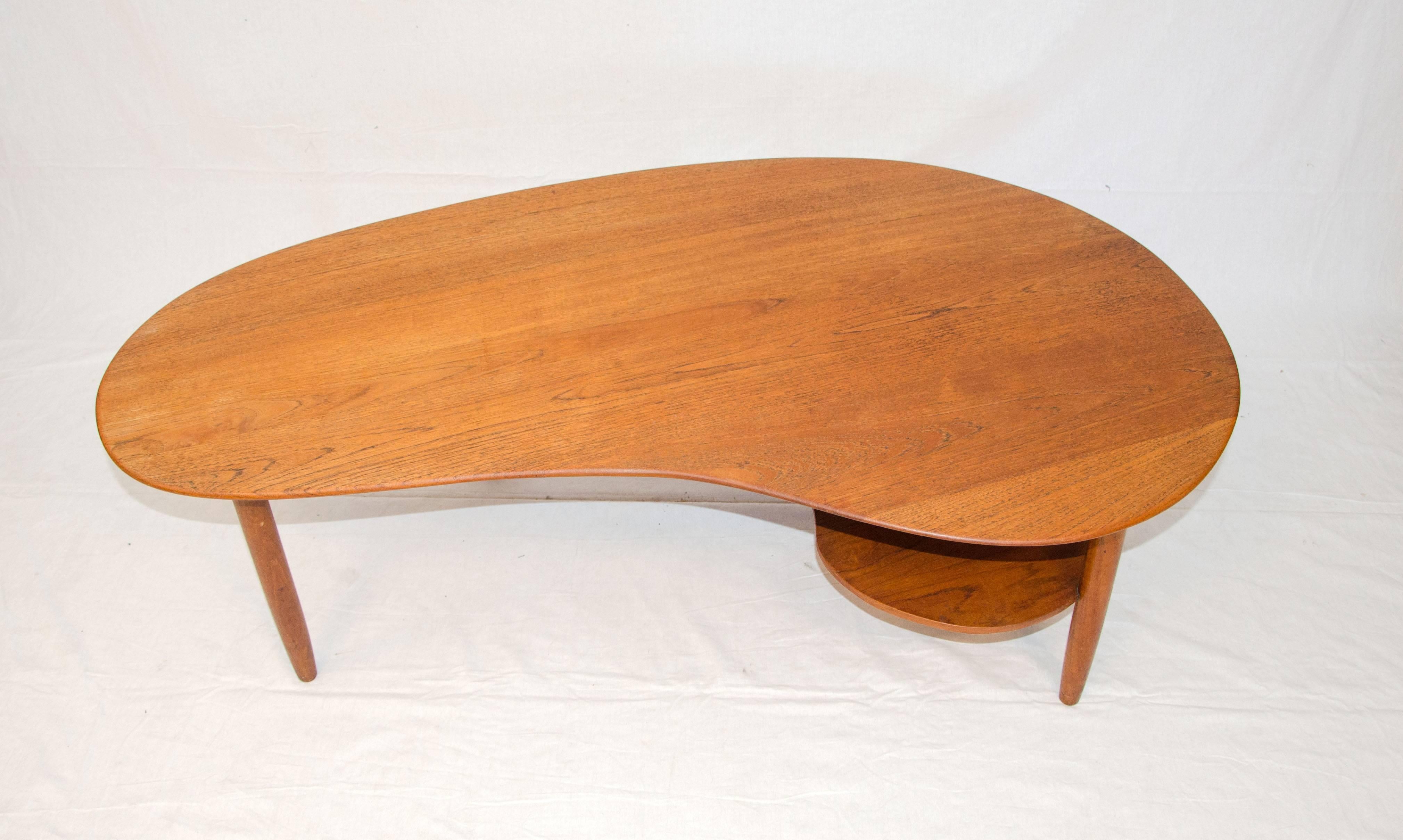 Nice teak kidney or organic shaped coffee table with a second smaller shelf under the top. The top and round tapered legs are solid teak. The second shelf is teak veneer. The second shelf is shaped somewhat like a painter's palette.
The second